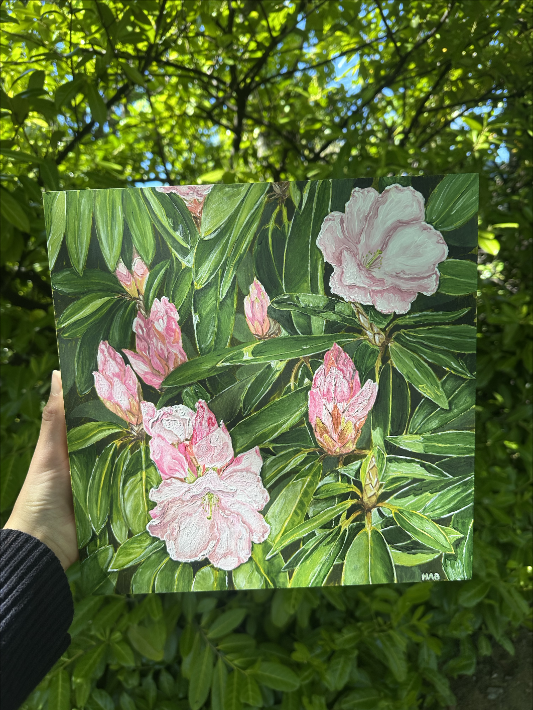A hand holding a painting of pink rhododendron flowers and green leaves, with a background of green foliage and blue sky.