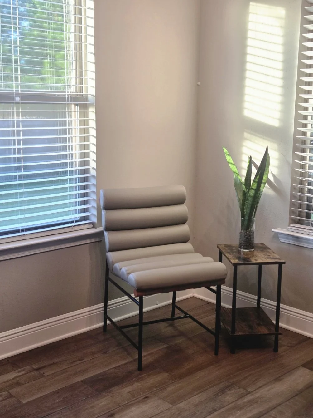 Modern chair with cylindrical cushions next to a small table with a potted snake plant in a glass vase, near two windows with blinds and natural sunlight.