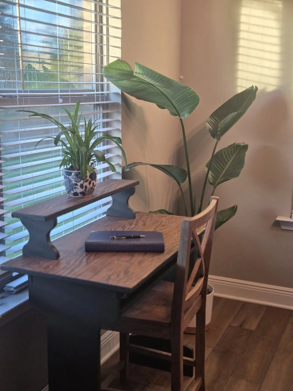 A wooden desk with a small potted plant on a raised shelf, a closed notebook, and a pen. Behind the desk, there are large green leafy plants near a window with white blinds, letting in natural light.
