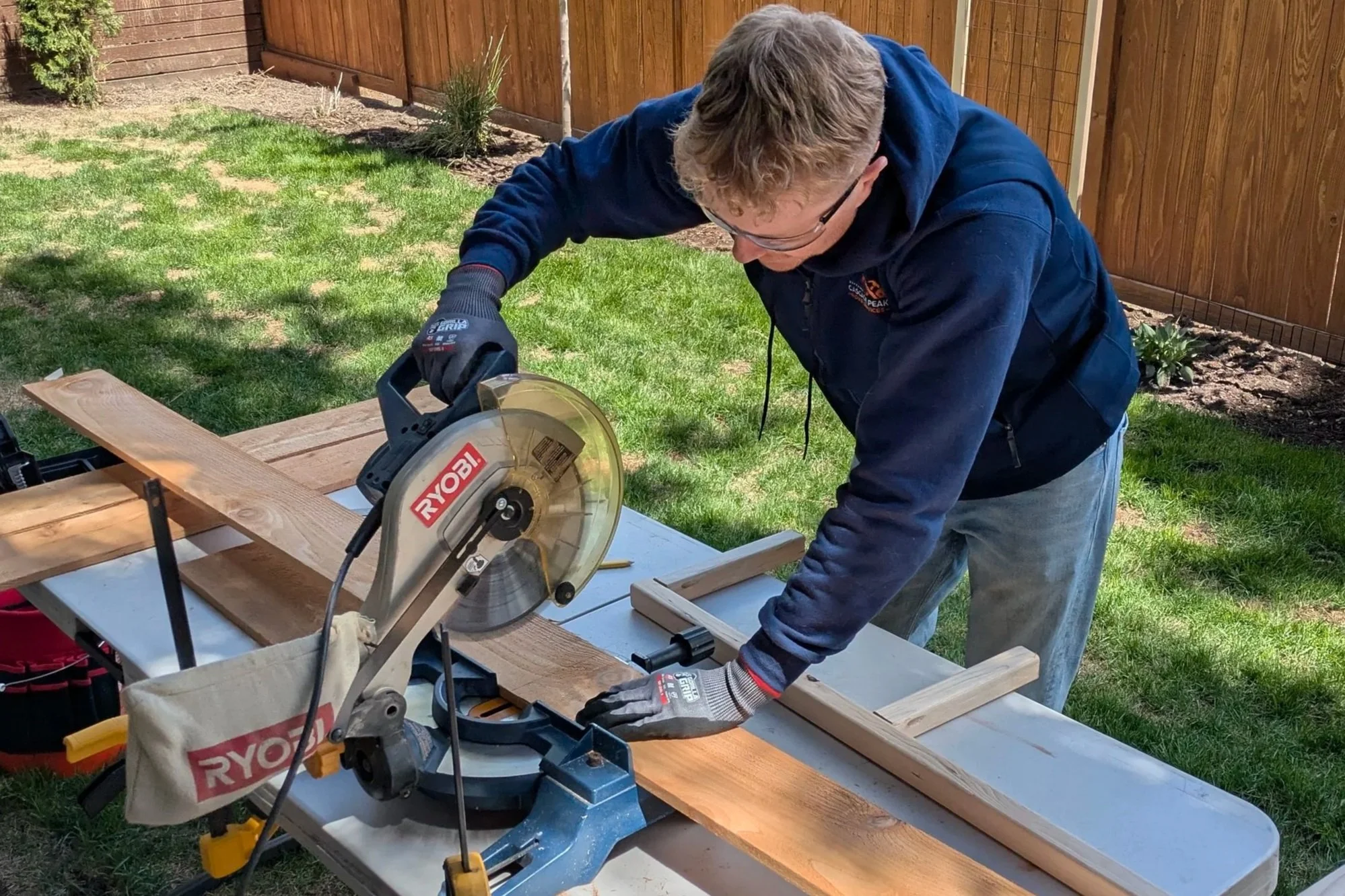 A man wearing glasses, gloves, and a blue hoodie is using a chop saw on a piece of wood outdoors. The scene shows a backyard with green grass, a wooden fence, and some small plants.