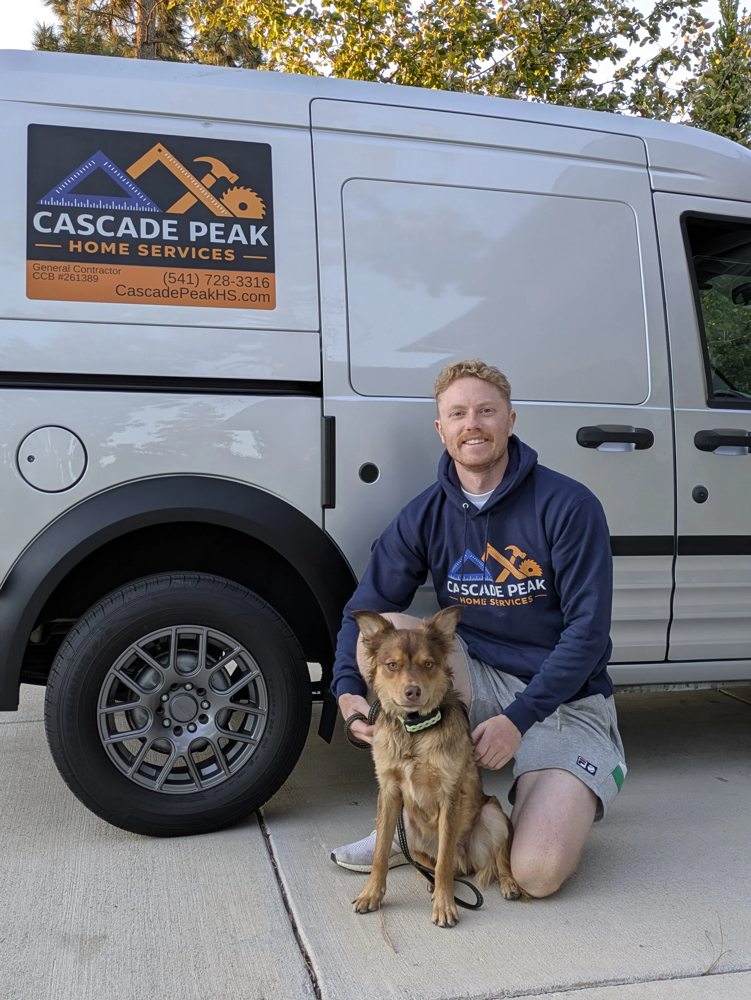 Man kneeling down with a happy dog in front of a white van with the logo for Cascade Peak Home Services. The man is wearing a hoodie with the company's logo. The dog is wearing a collar and looks at the camera. The van has a ladder and tools painted on its side.