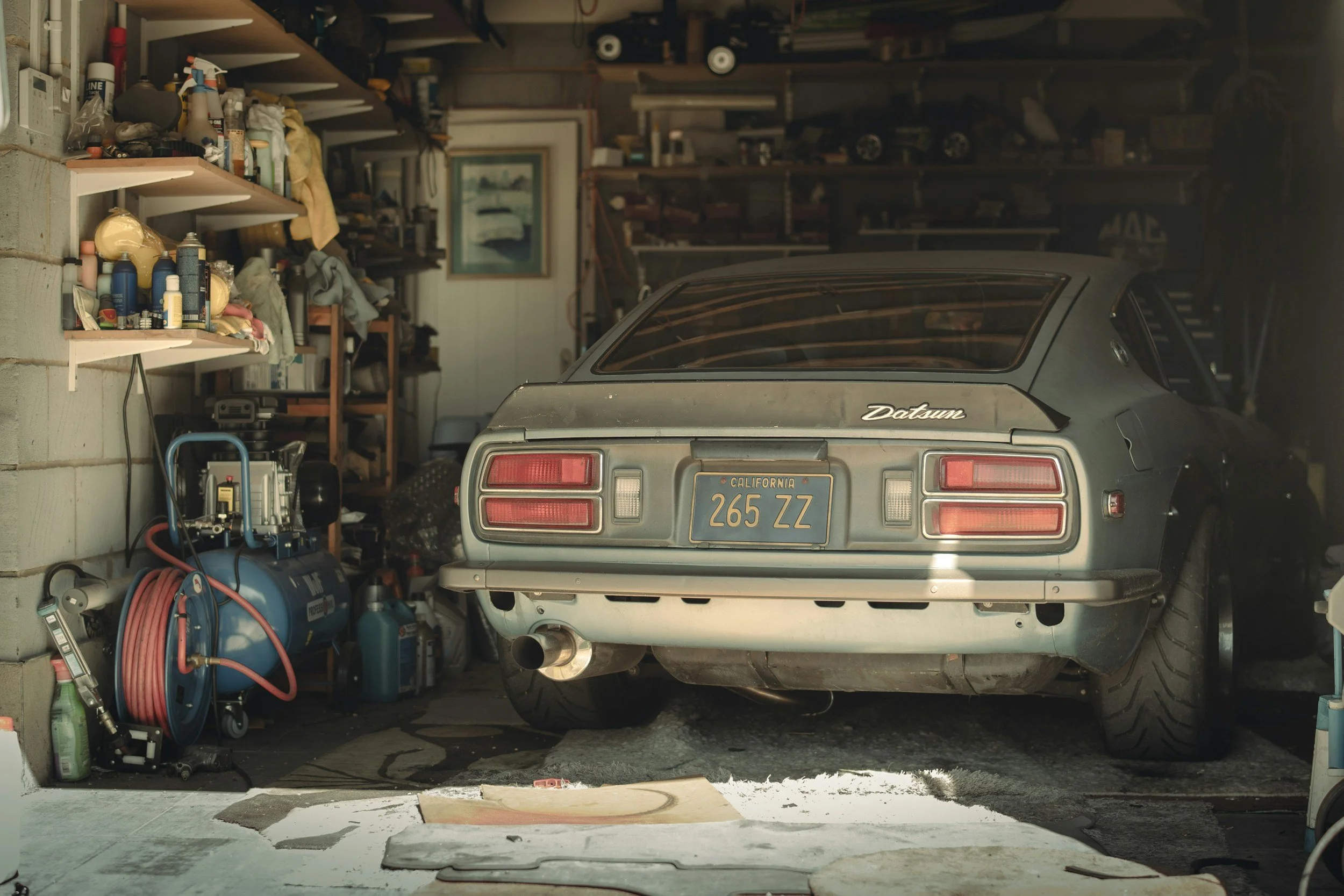 A garage with shelves filled with various cans, bottles, and tools on the left, and a vintage gray Datsun car with California license plate 265 ZZ parked inside. The garage has a concrete floor, some tools, and a painting on the back wall.