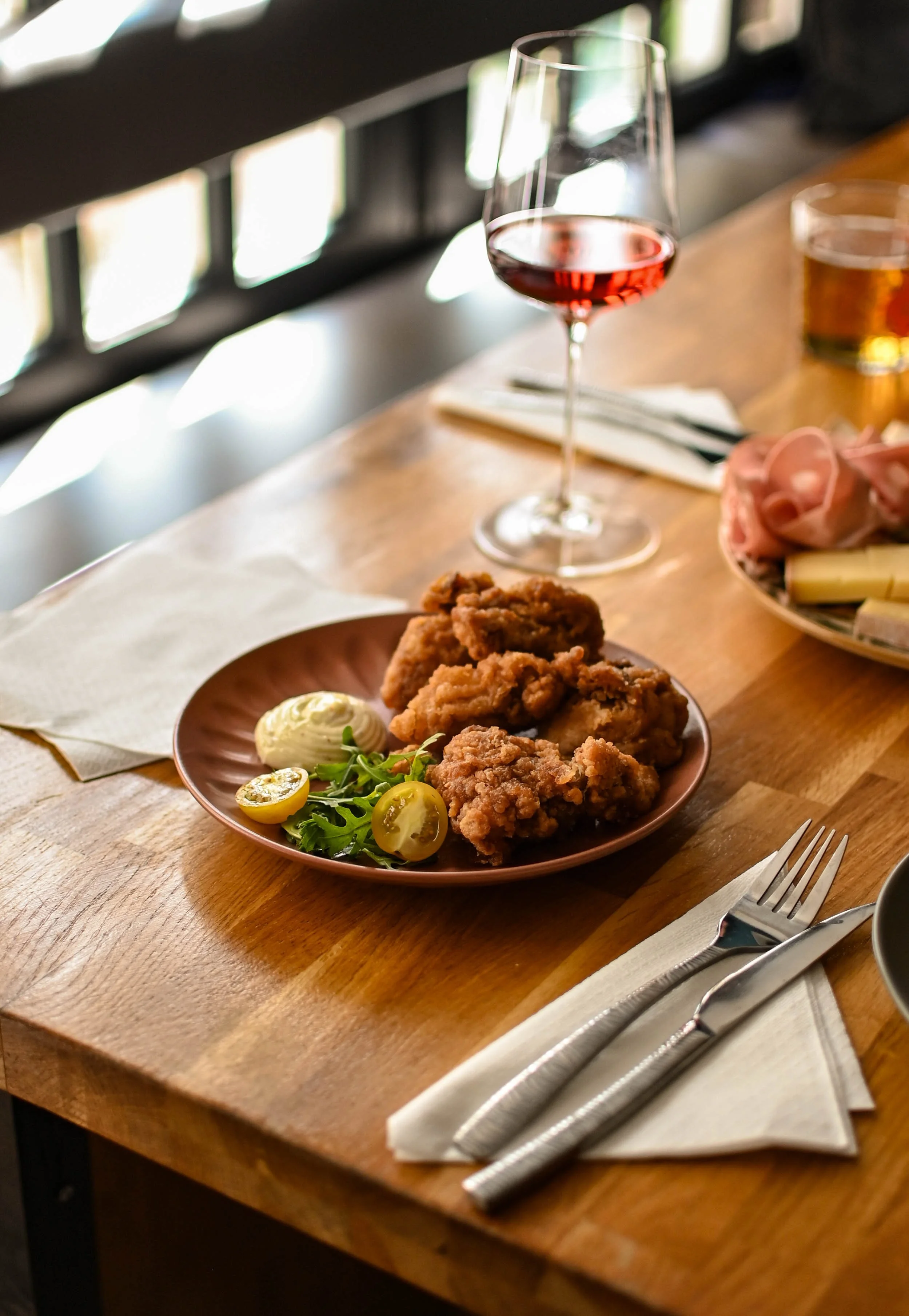 Une assiette de poulet frit accompagnée de salade verte, de tomates cerises et de mayonnaise, une coupelle de bière, un verre de vin rosé, et une assiette de charcuterie et de fromage sur une table en bois avec des couverts en metal et un linge blanc