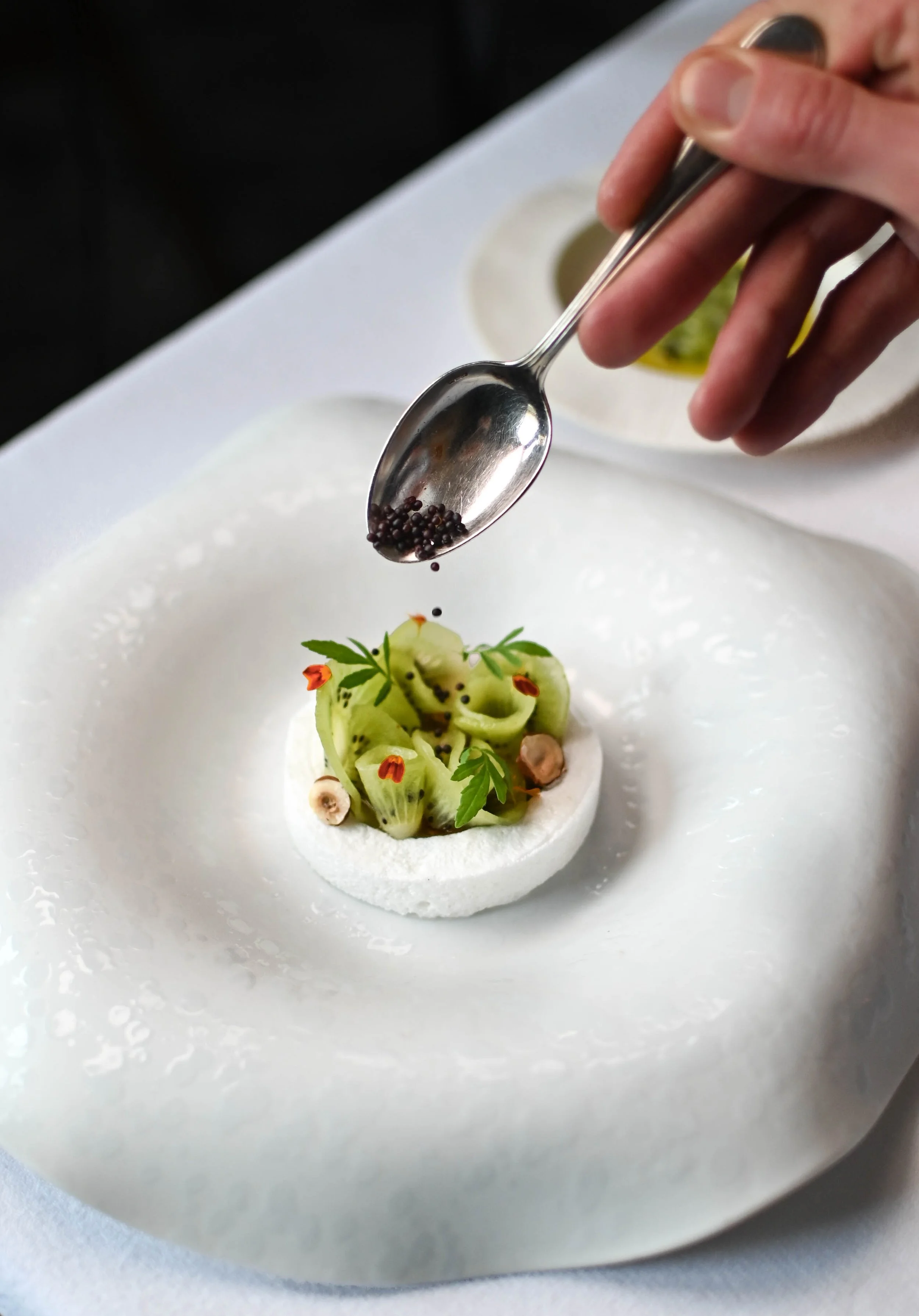 Un dessert élégant avec des tranches de kiwi, des fleurs comestibles, et une garniture de graines de colza, présenté dans un cercle de mousse blanche sur une assiette blanche.