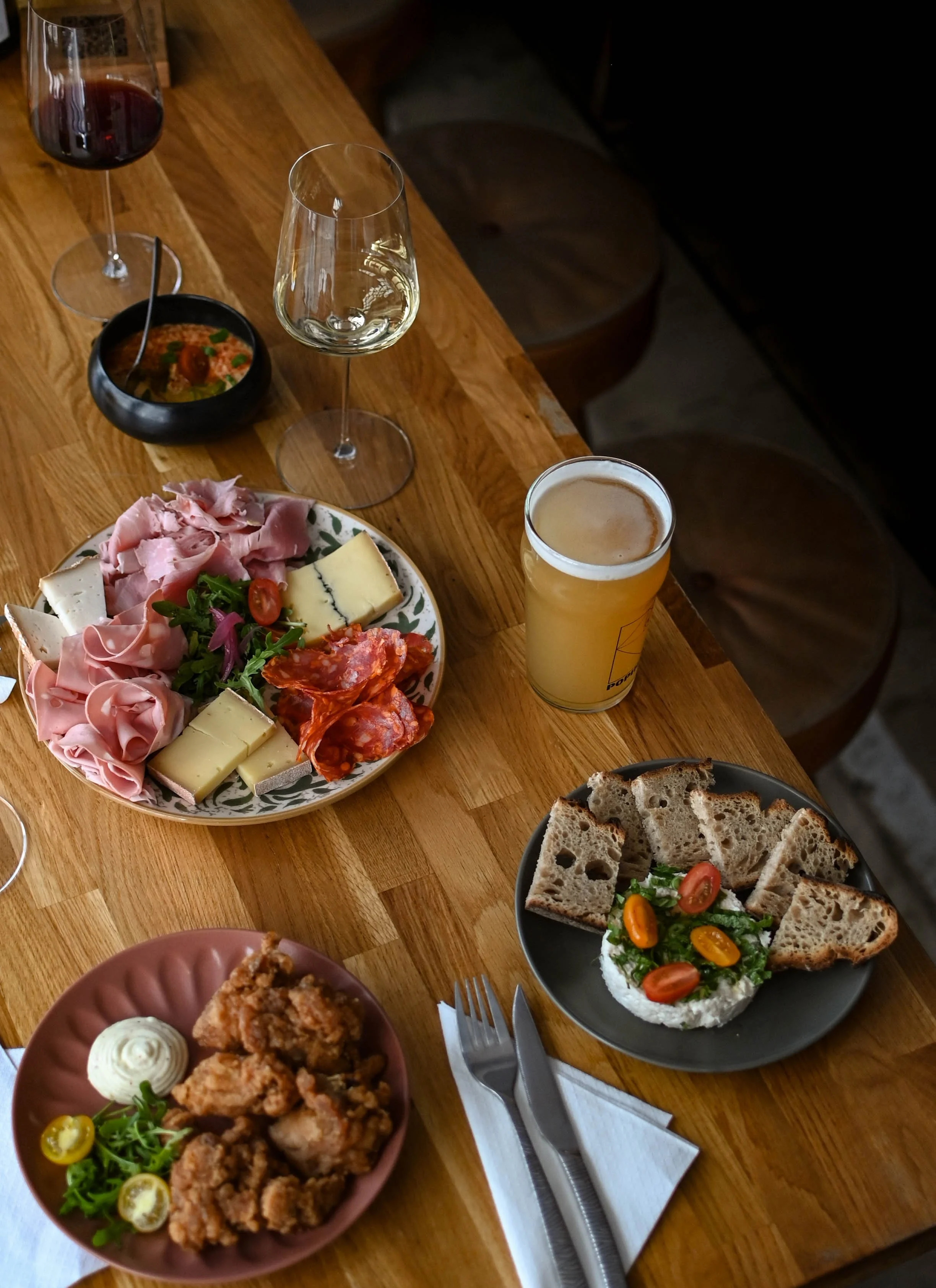 Plate de charcuterie avec jambon, fromage, tomates cerises, salade, pain, bière, verres de vin blanc et rouge, et différentes assiettes avec des amuse-gueules.