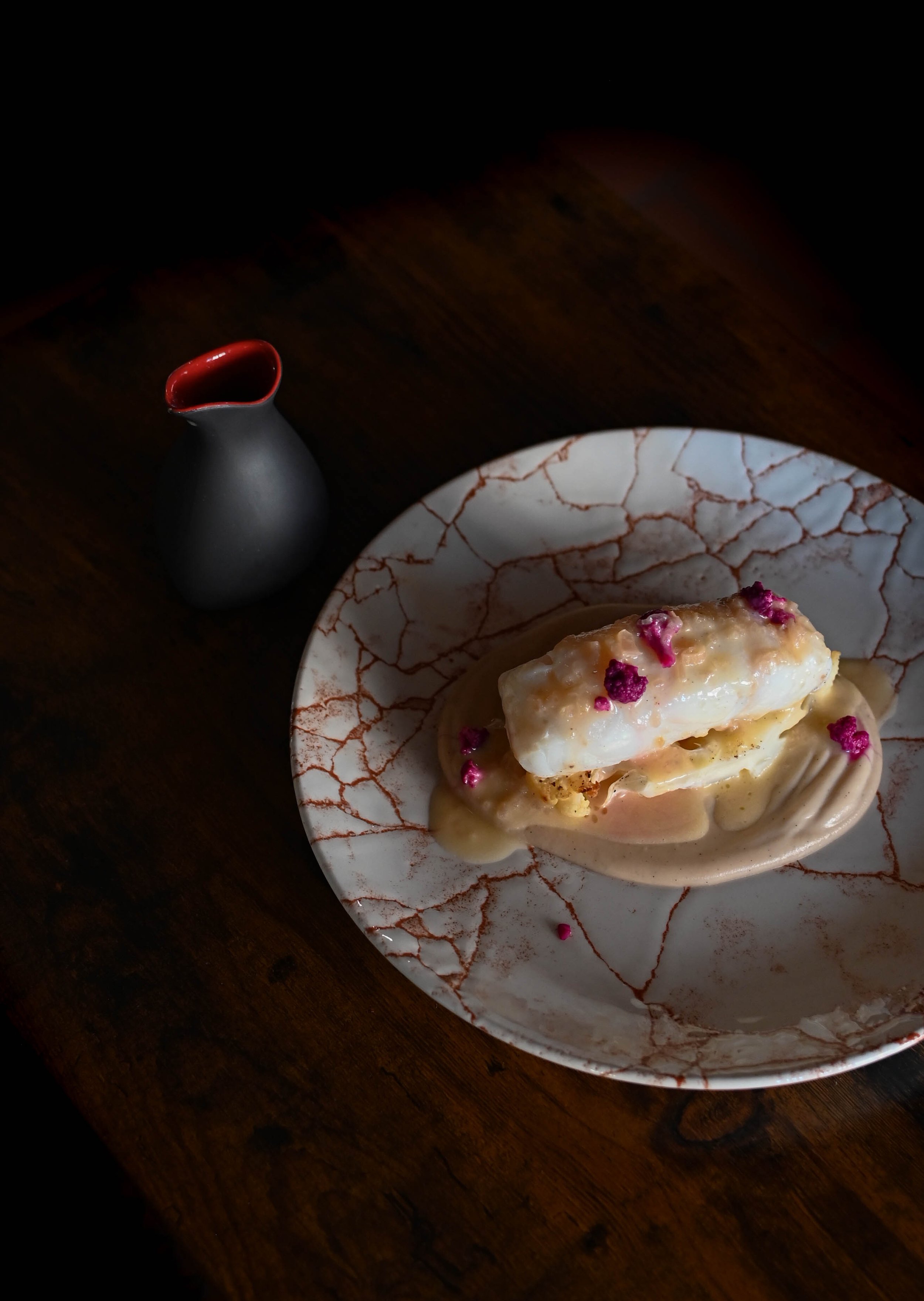 Une patisserie avec une couche de crème, décorée de pétales de fleurs violettes sur une assiette marbrée blanche et orange, accompagnée d'une petite cruche noire avec intérieur rouge, sur une table en bois.