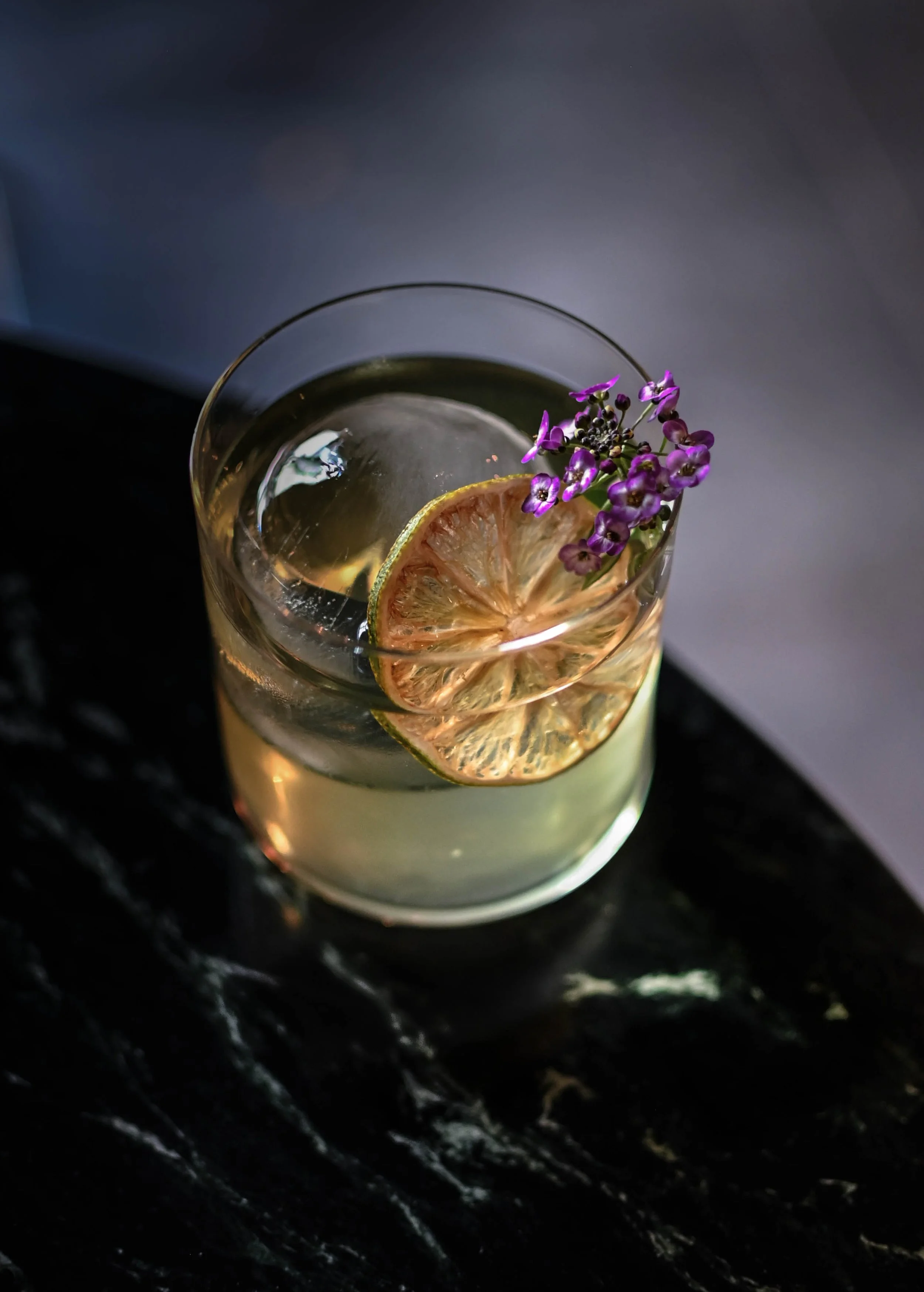 Un verre contenant une boisson claire avec une tranche de citron et des petites fleurs violettes en garniture, posé sur une surface sombre.