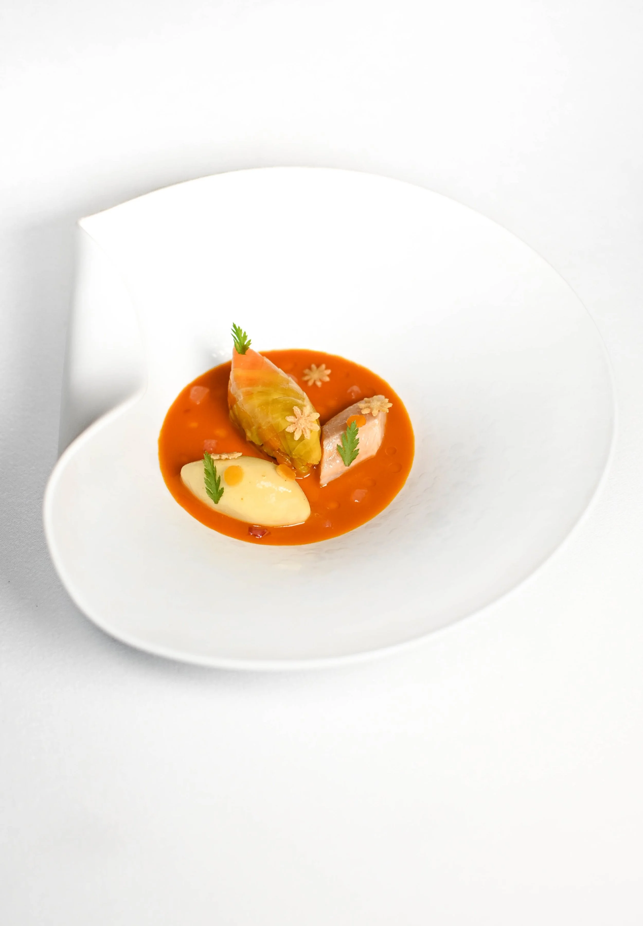 Plat blanc avec un saucisson enroulé dans une feuille de chou, accompagné d'une sauce rouge, garnitures de petites fleurs et de jeunes pousses de verdure.