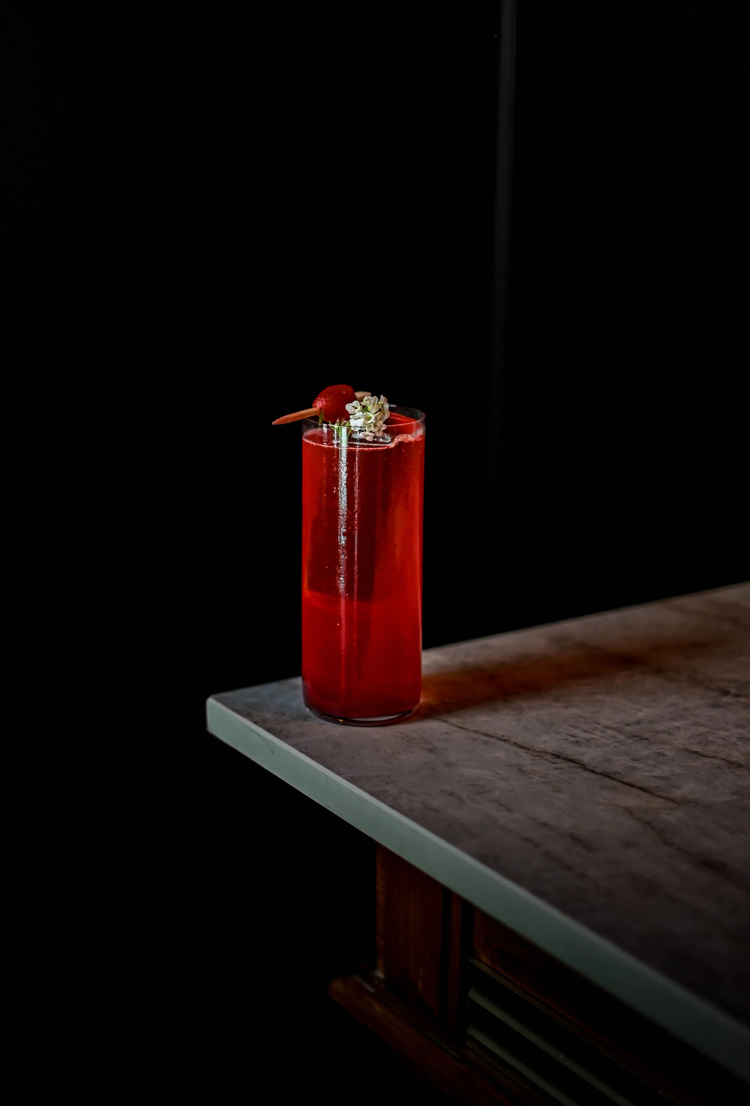 Un cocktail rouge dans un verre haut placé sur un comptoir en bois, décoré d'une cerise, d'une petite branche de fleurs blanches et d'une tige de fraise, avec un fond noir.