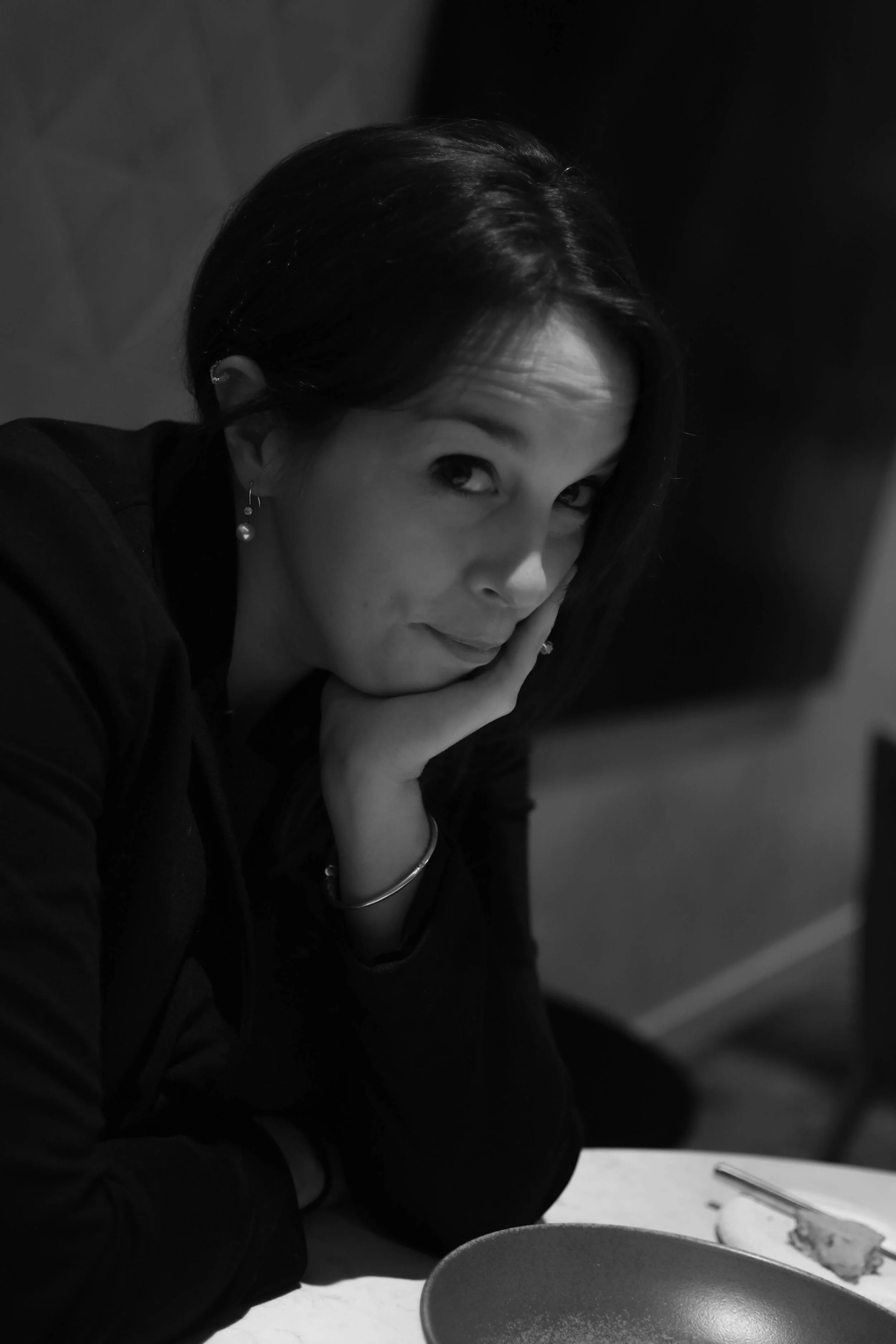 Une femme aux cheveux noirs et courts, portant des boucles d'oreilles pendantes, est assise à une table. Elle a la main sur le visage et regarde vers l'objectif. Elle semble être dans une cuisine, avec une assiette de pizza sur la table.