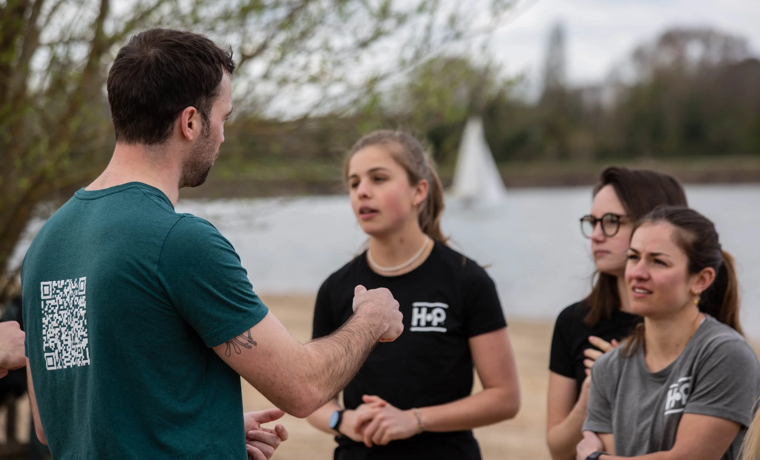 Groupe de jeunes femmes écoutant un homme en conversation dehors près d'une rivière, arrière-plan avec arbres et bateau à voile.