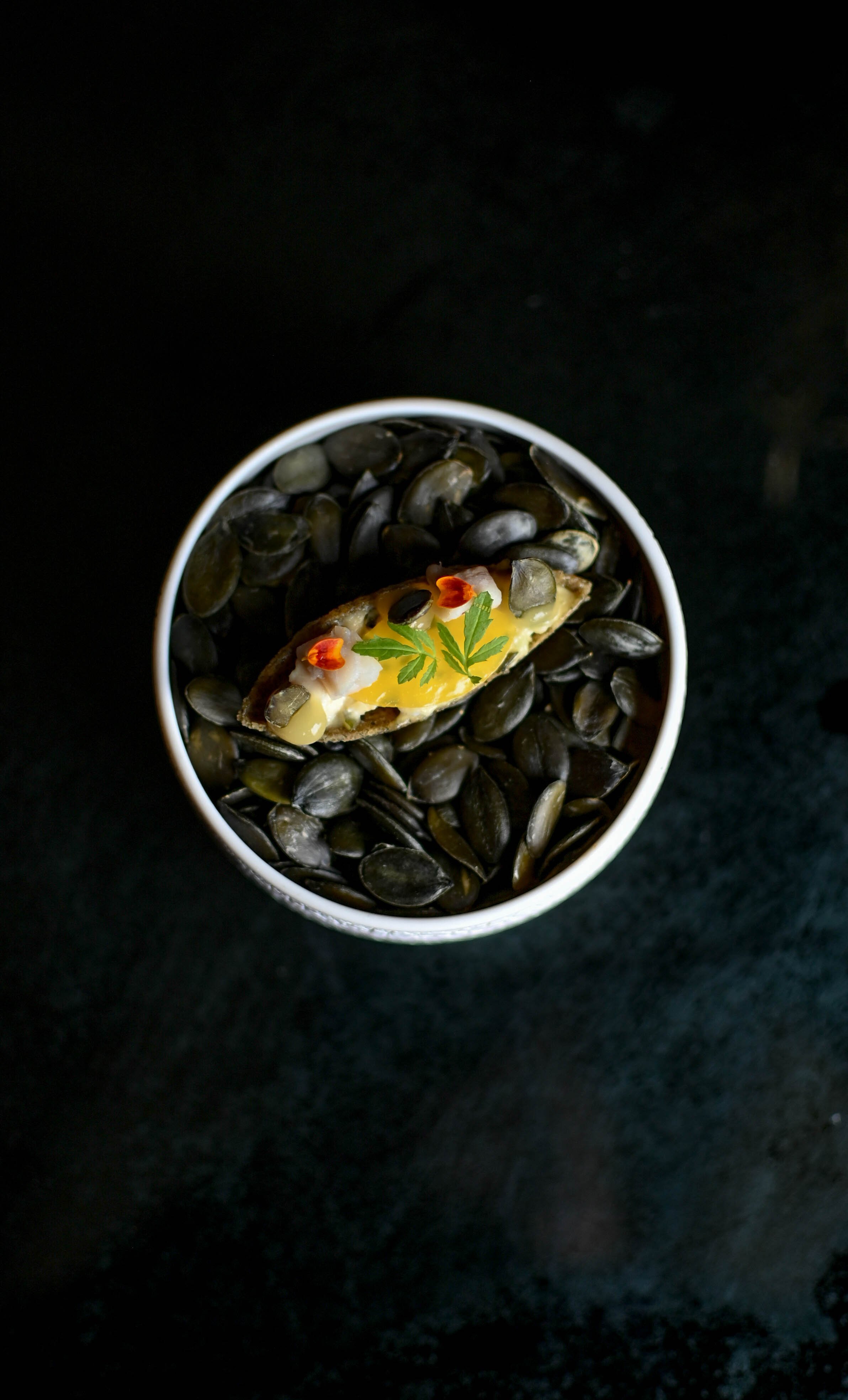 Un bol rempli de graines de citrouille, avec une tranche de citron, une petite feuille verte, et quelques pétales de fleurs rouges