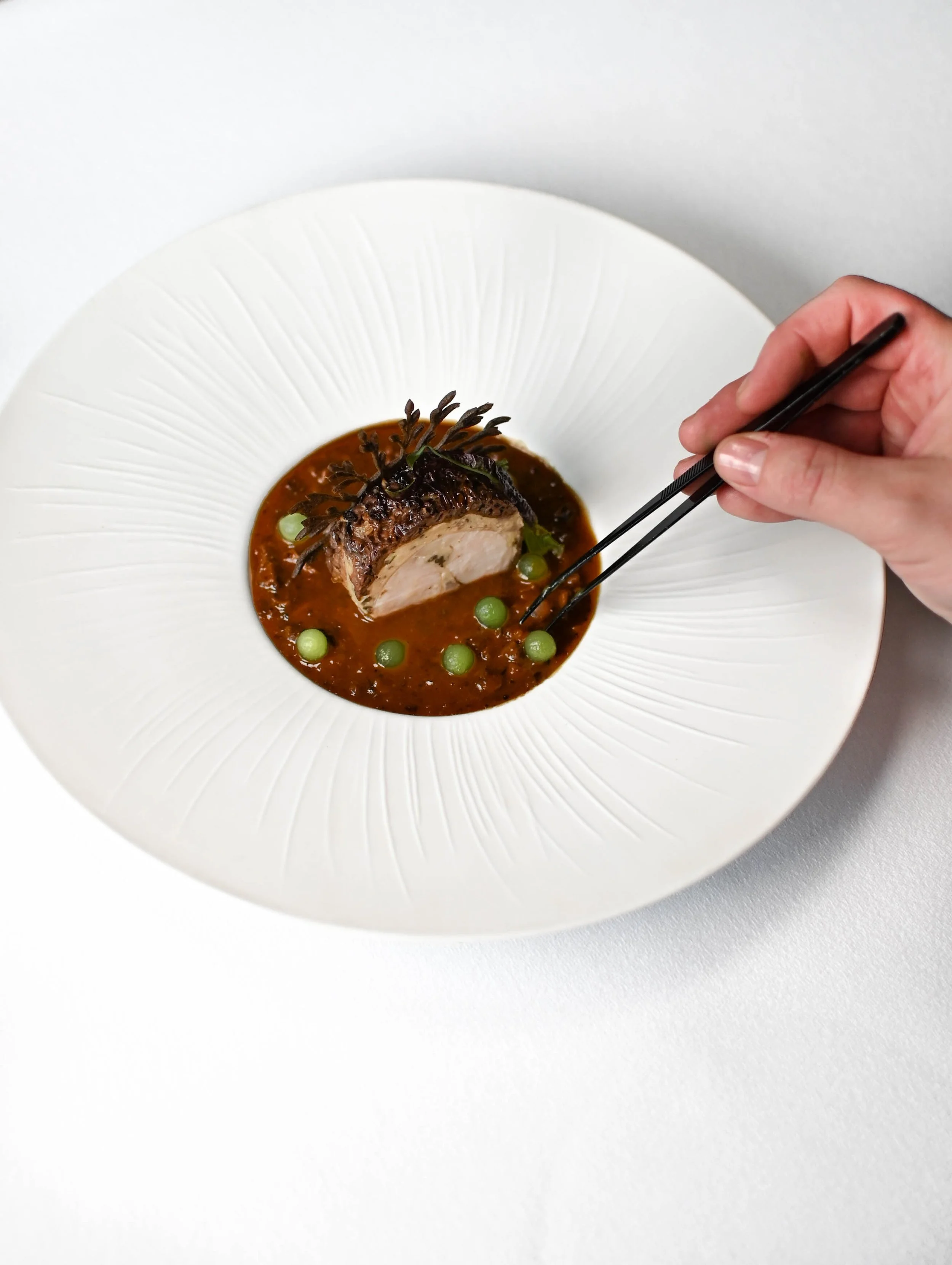 Plat blanc avec un morceau de viande, sauce, petits pois verts, et garnitures végétales, avec des baguettes japonaises