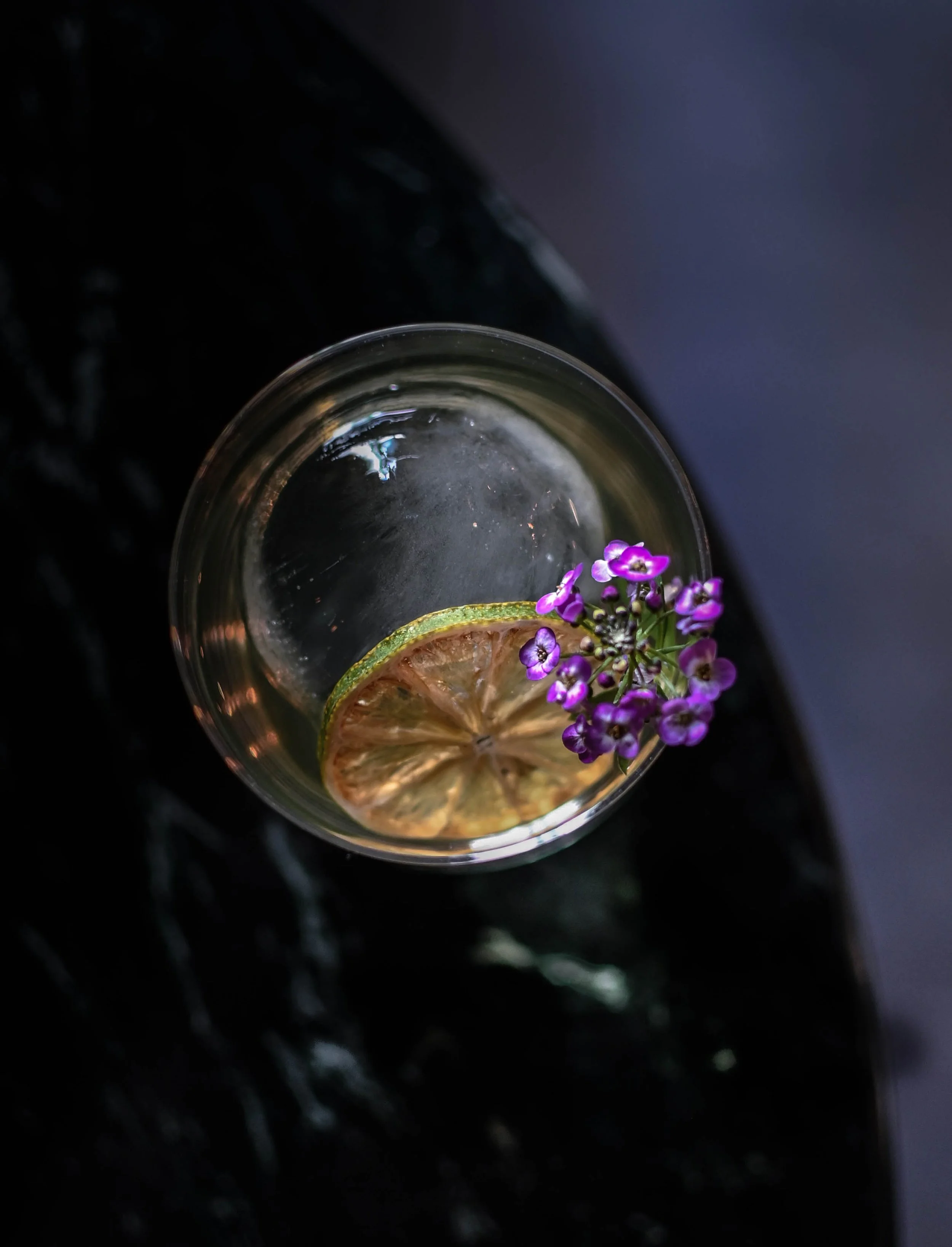 Verre transparent rempli d'une boisson claire avec une tranche de citron et des petites fleurs violettes en décoration.