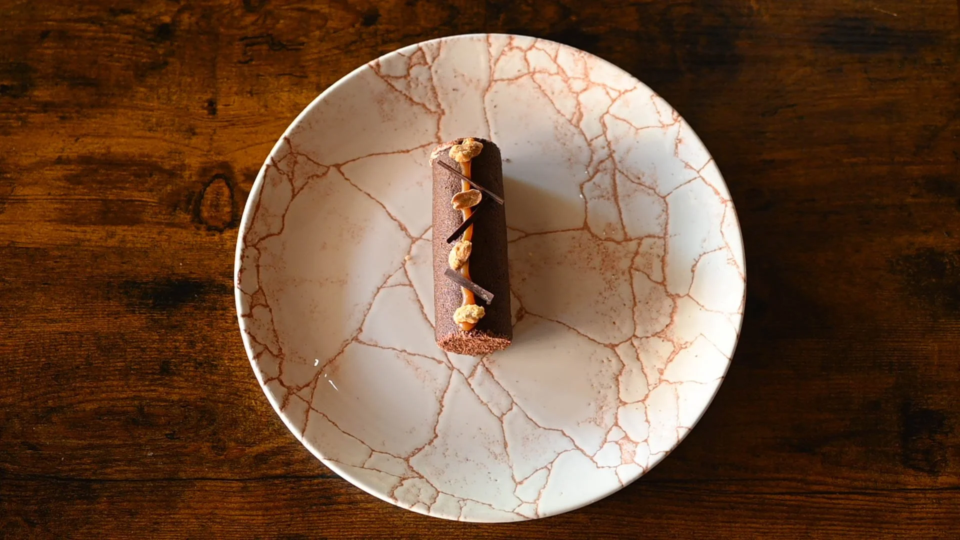 Un dessert au chocolat posé sur une assiette blanche avec un motif de fissures marron, placé sur une table en bois.