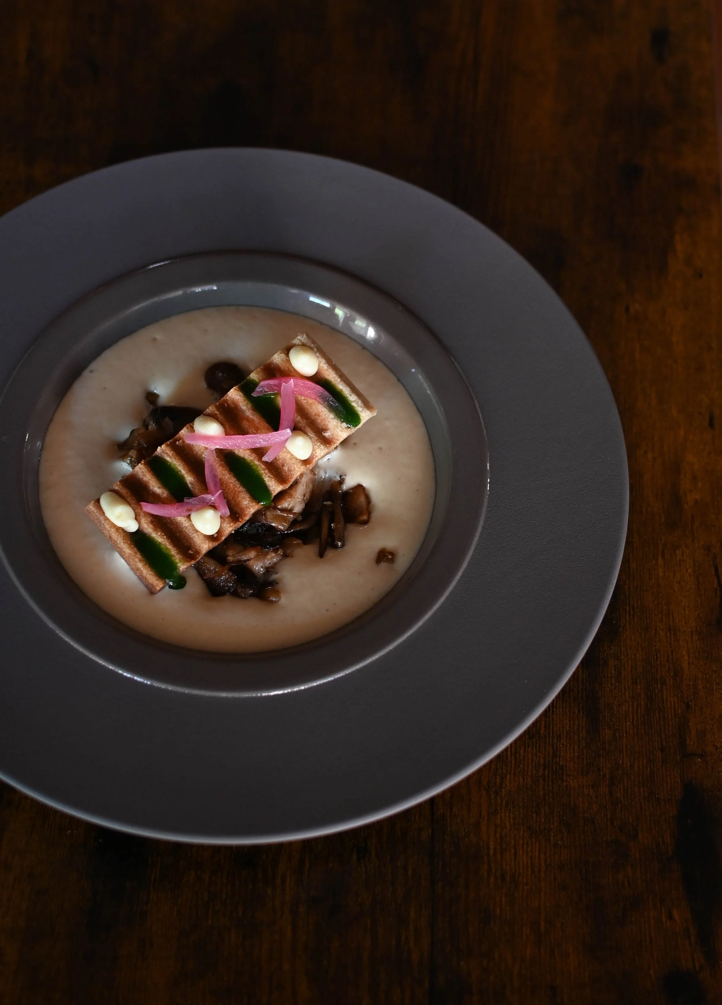 Soupe ou crème dans un bol gris, garnie d'un morceau de gâteau ou de biscuit décoré de crème, de fines tranches d'oignon et de sauce verte, avec un décor en fond noir.
