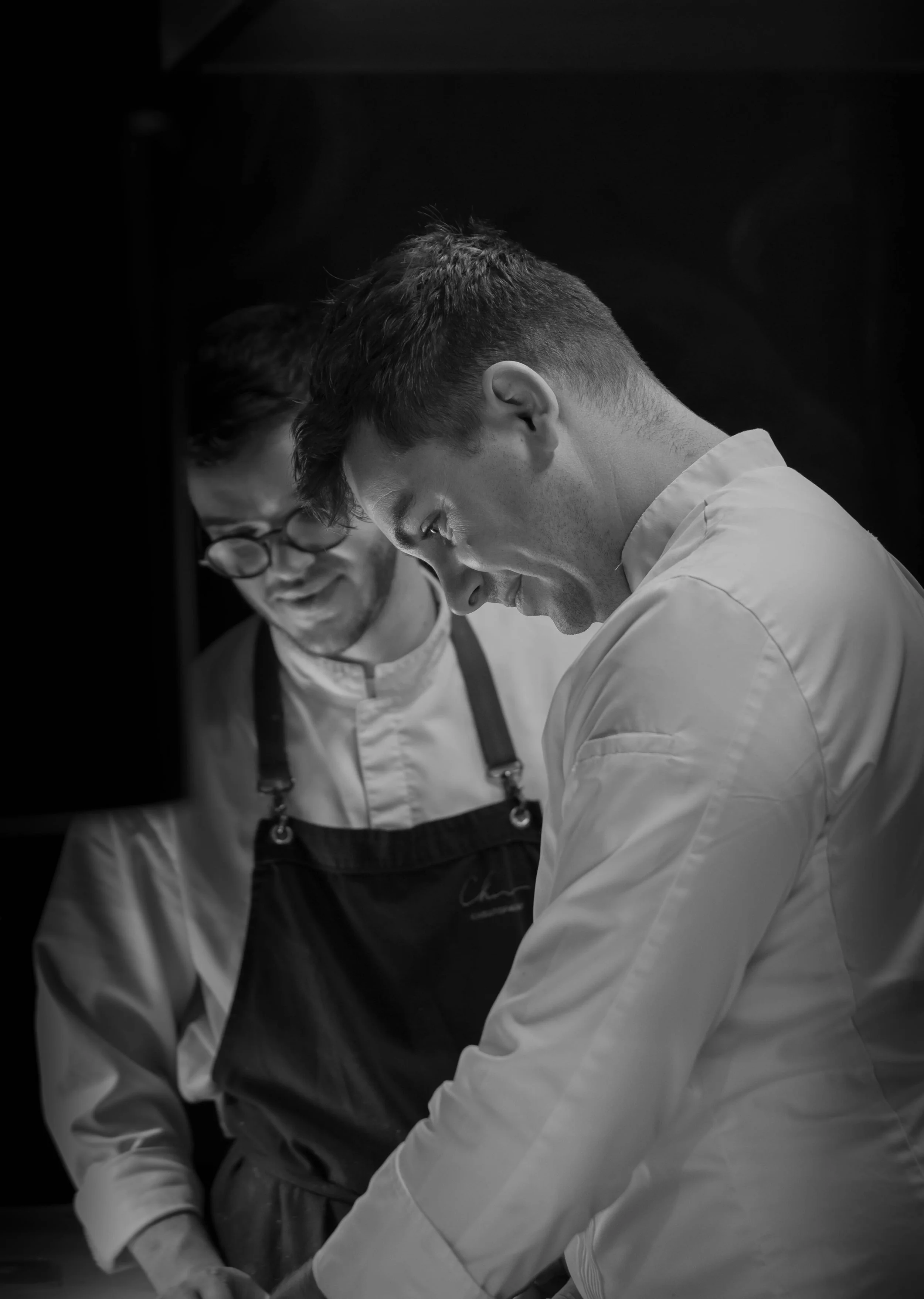 Deux chefs de cuisine en train de préparer un plat dans une cuisine. L'un porte une veste blanche et un tablier noir, l'autre porte une veste blanche.