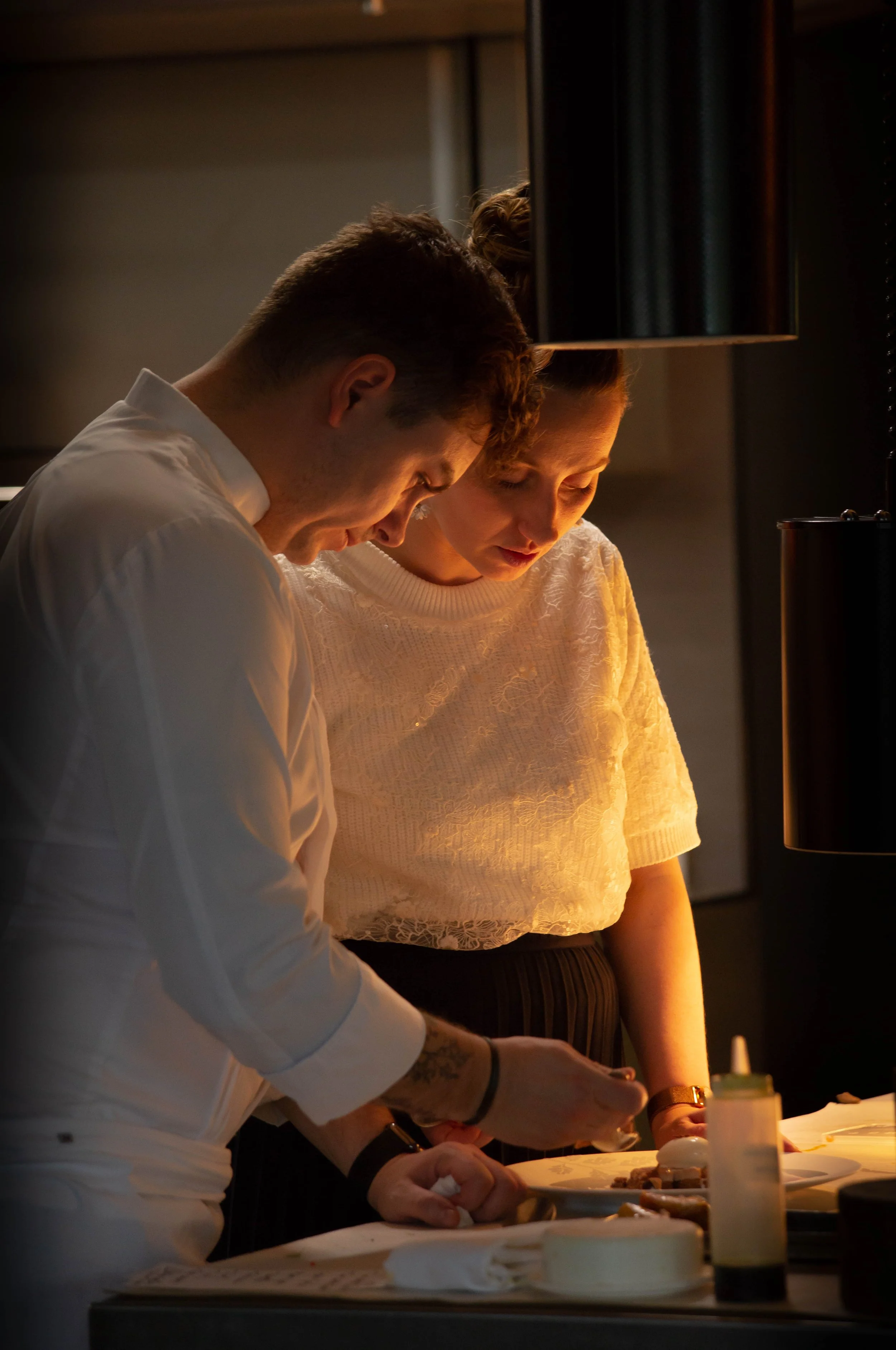 Deux personnes préparent des plats dans une cuisine, éclairées par une lumière chaude, avec des ingrédients sur la table.