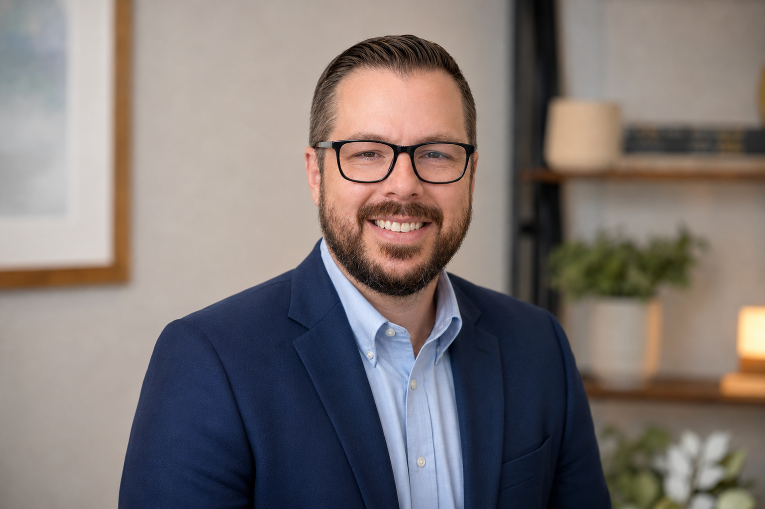 A man with glasses, brown hair, and a beard smiling, wearing a blue suit jacket and light blue shirt, sitting in an office with shelves, plants, and framed artwork in the background.