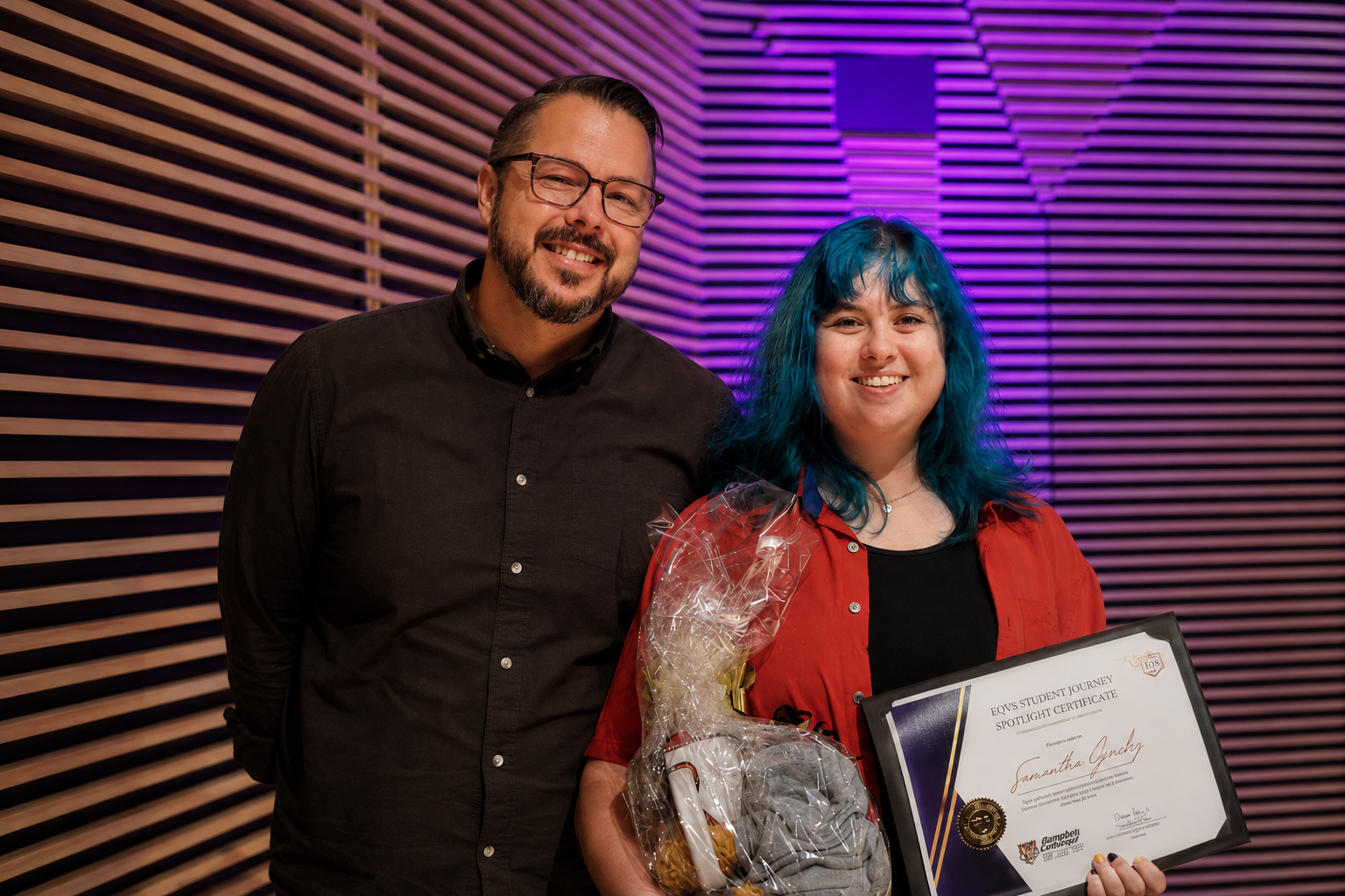 A woman with blue hair holding a certificate and a gift bag, standing next to a man in a black shirt, in a room with purple and wooden wall decor, both smiling at the camera.
