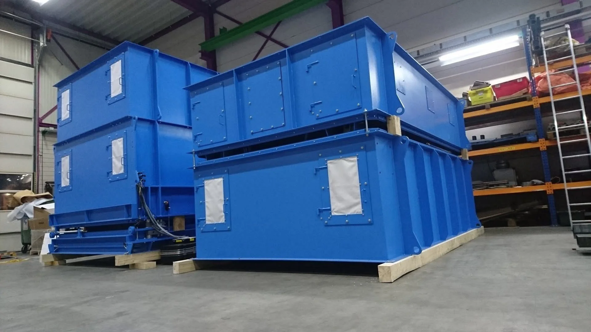 Grote blauwe technische apparatuur in een magazijn, op houten platformen, met opslagrekken op de achtergrond.