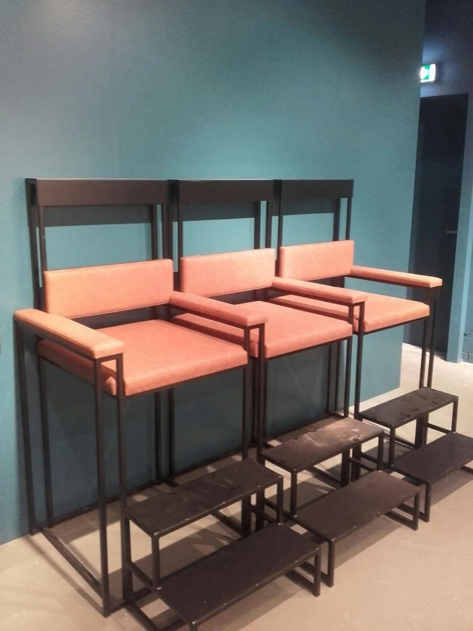 Drie beige zitbanken op hoge metalen frames hangen aan een blauwe muur, met drie zwarte stoeplasten eronder.