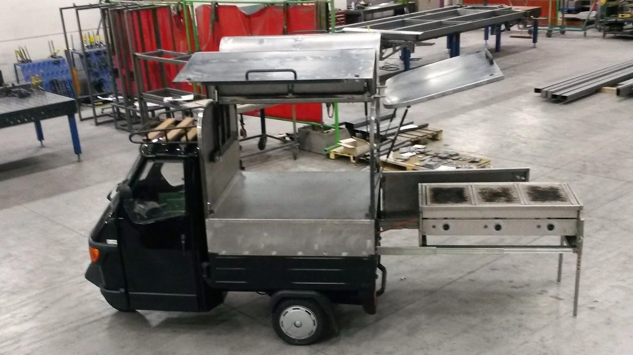 Een kleine zwartgebouwde vrachtwagen met een open laadbak die vol met metalen voorwerpen staat, binnen een magazijn of werkplaats met metalen stellingen en gereedschap op de achtergrond.