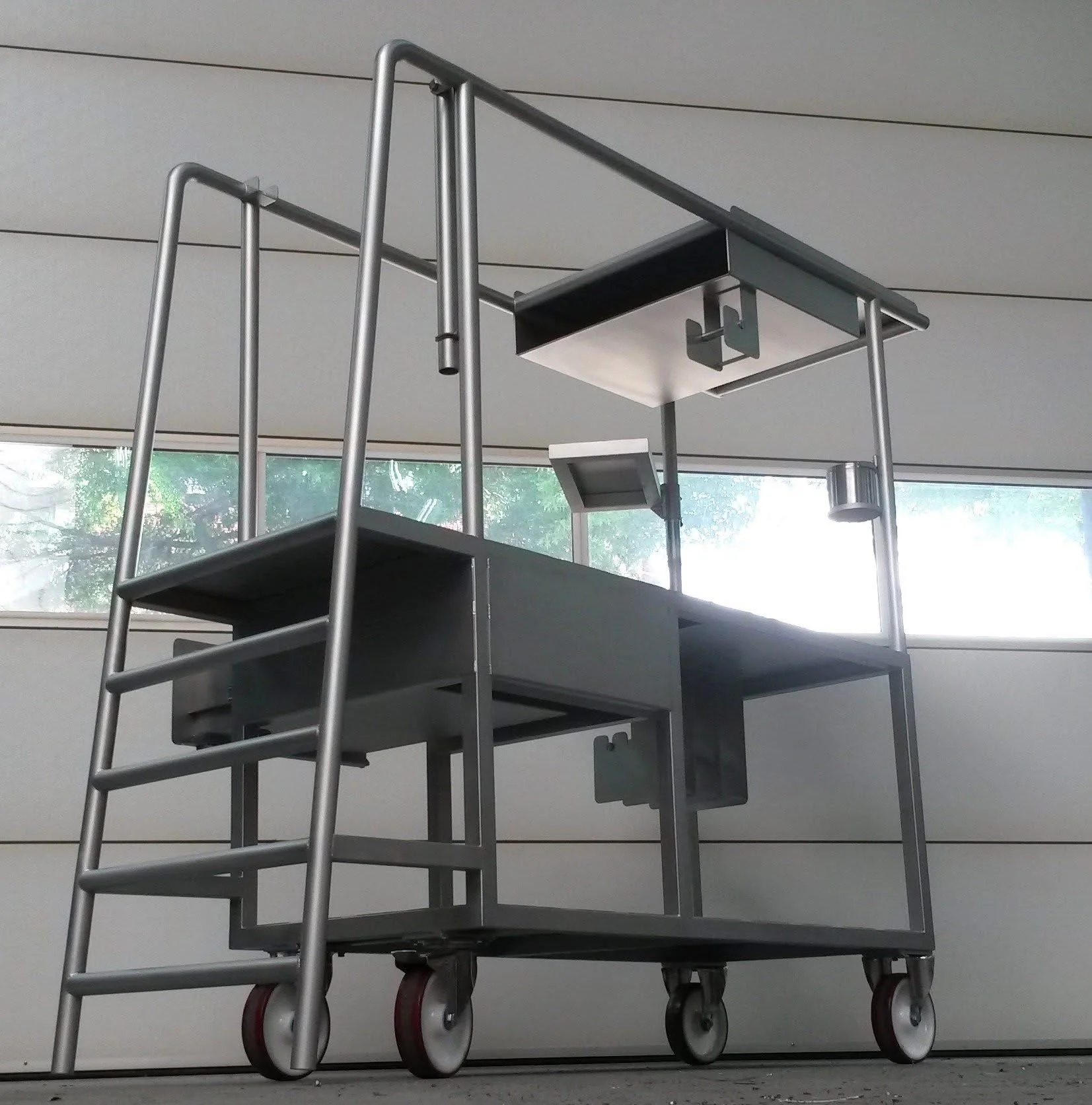 Stalen karretje met verschillende onderdelen en een tafel of robotarm, geplaatst in een binnenruimte met grote ramen en een plafond met horizontale lijnen.