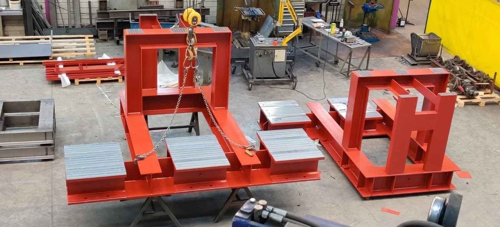Twee grote rode metalen constructies in een werkplaats, met sommige onderdelen en gereedschappen eromheen.