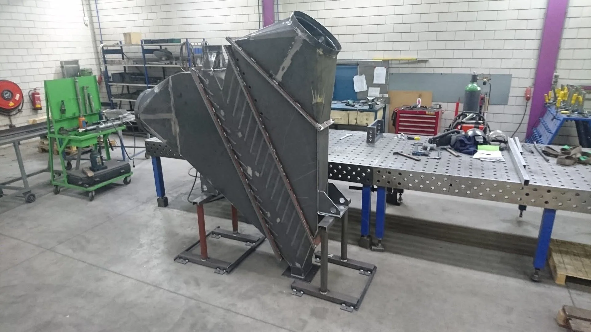 Werkbank met metalen onderdelen in een werkplaats, omgeven door gereedschap en apparatuur.
