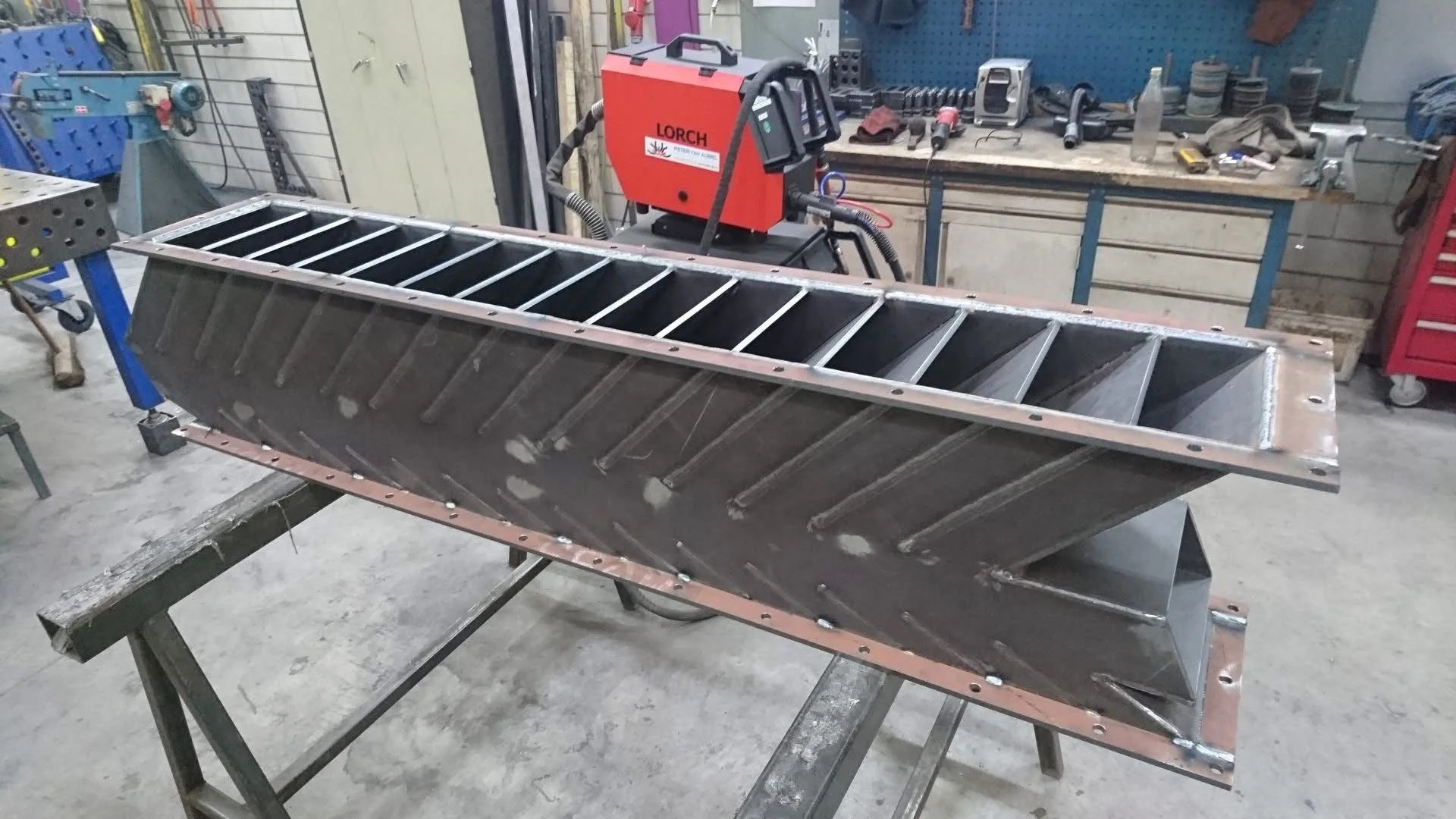 Metaalconstructie met interne structuur en lasnaden, in een werkplaats met gereedschap en werkbanken.
