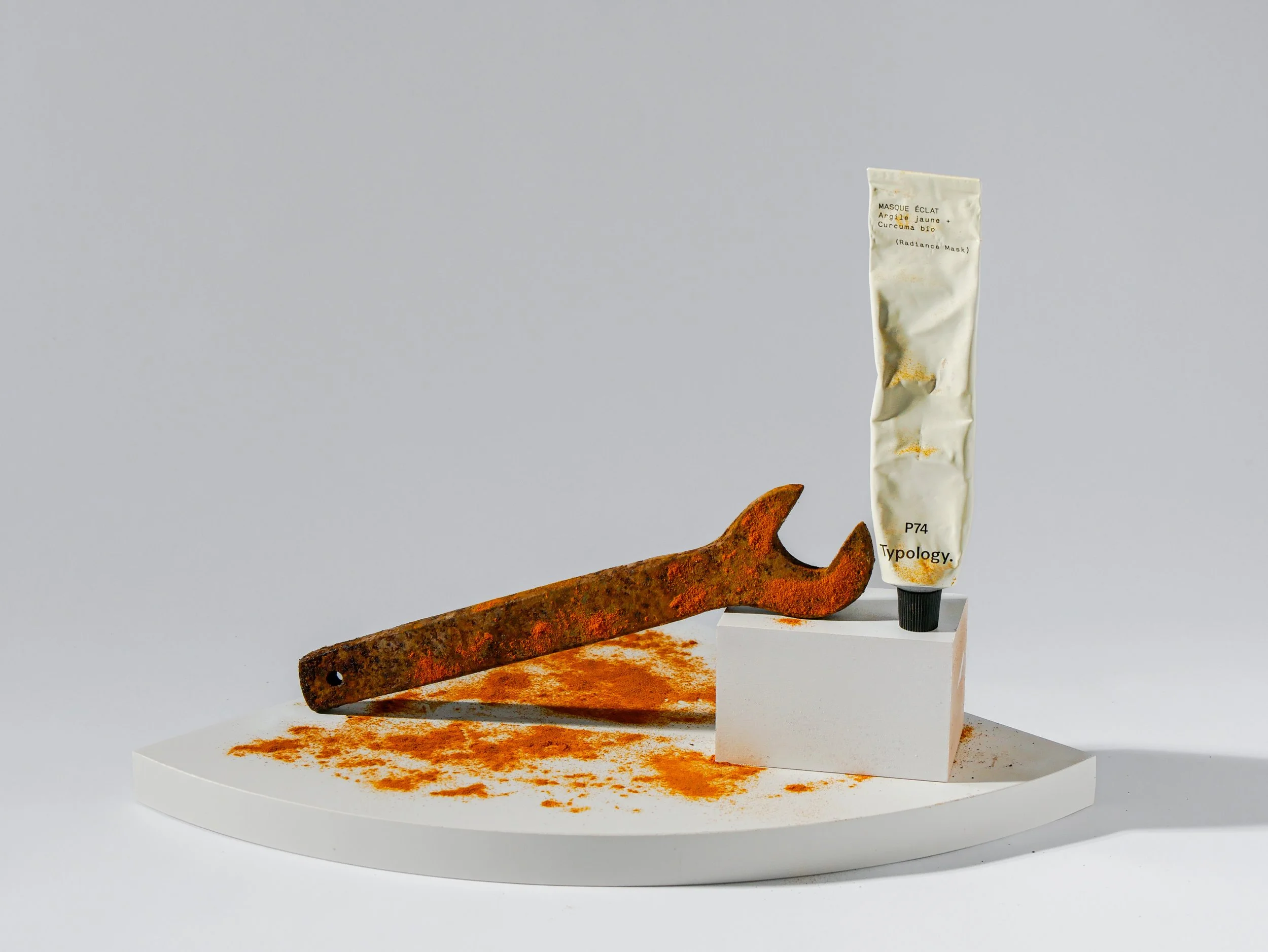 An art installation featuring an old rusty iron piece, a tube labeled "Radiance Mask" with yellowish-orange powder, and an orange powder scattered on a white surface, all arranged on a white oval base.