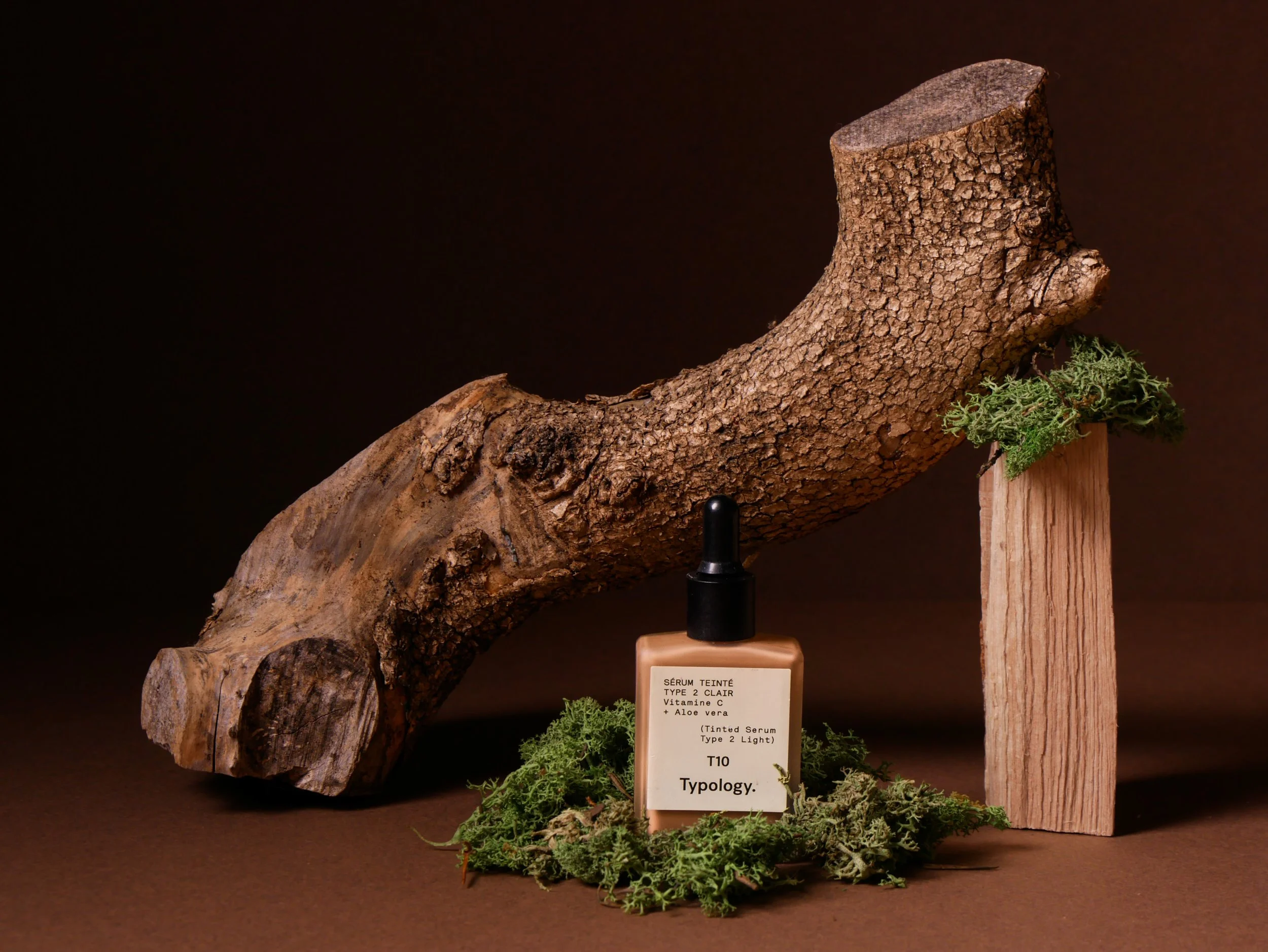 A wooden display features a large piece of bark with a label that reads 'Serum Tinted Type 2 Clair, Vitamin C + Aloe Vera, T10. Typology.' Green moss surrounds the base of the display, and a small black dropper is positioned in front of the bark.