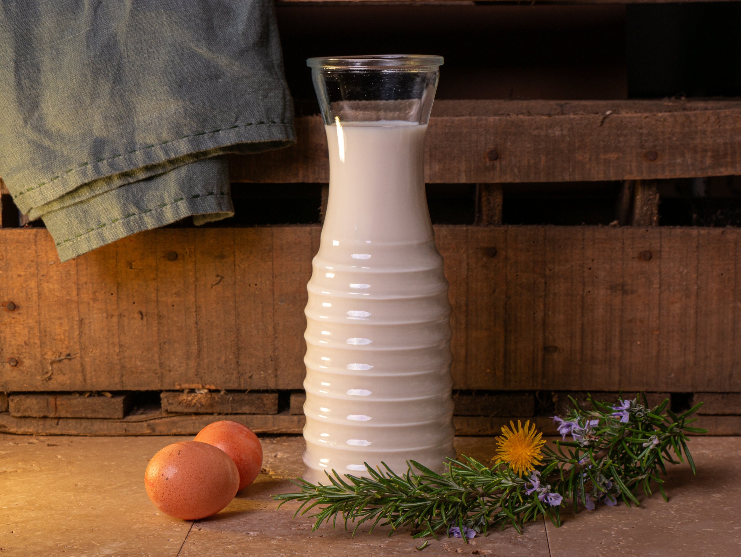 A white ceramic vase with a wavy pattern, two brown eggs, and a sprig of greenery with purple and yellow flowers on a wooden surface against a wooden background.