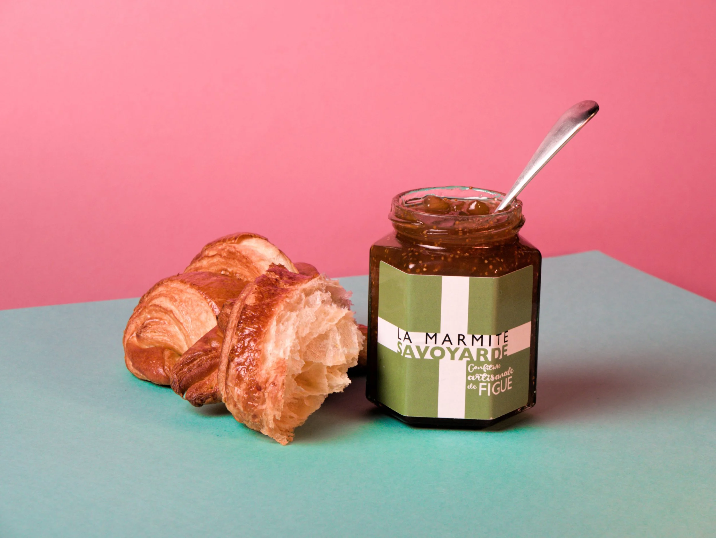 A jar of fig jam with a silver spoon inside, accompanied by sliced croissants on a pastel blue surface with a pink background.