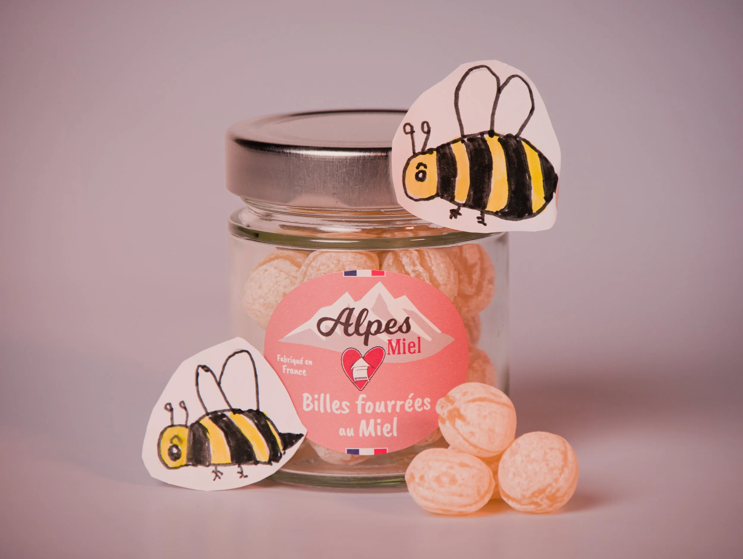 Close-up of a jar with a metal lid, decorated with a illustrated bee sticker and a pink label that reads 'Alpes Miel' and 'Billes Fourrées au Miel'. The jar contains round, orange and white candies.