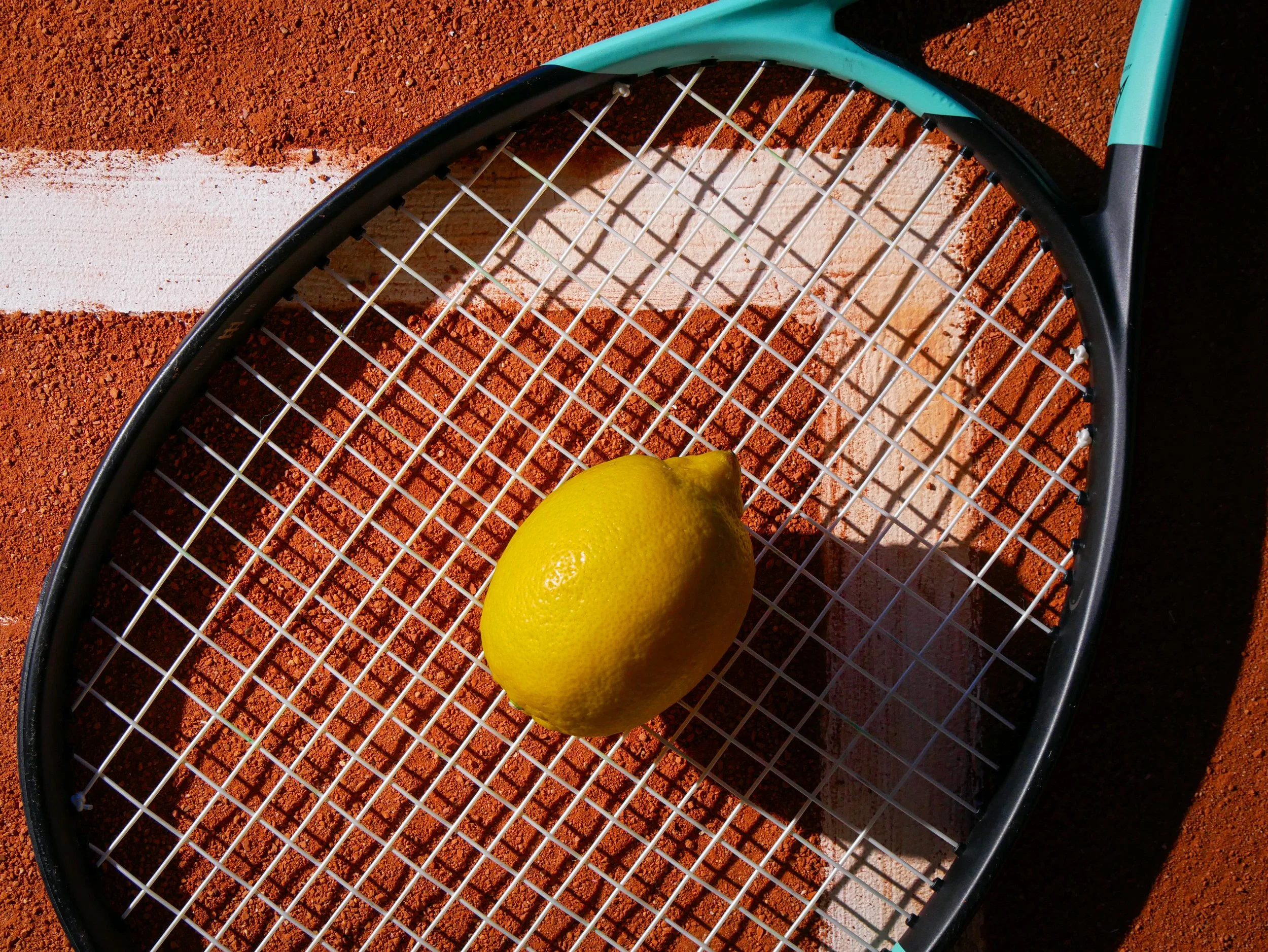 A tennis racket with a yellow tennis ball resting on it, placed on a clay tennis court with a white line.