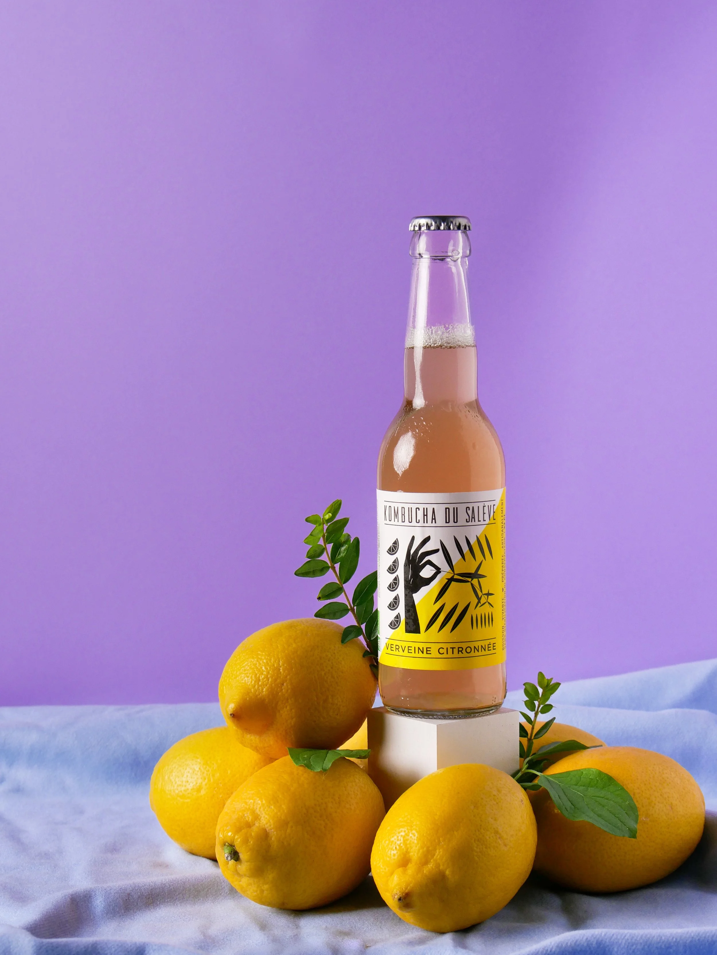 A tall bottle of kombucha labeled 'Kombucha du Salève Verveine Citronnée' is surrounded by fresh lemons and green leaves, set against a light purple background.