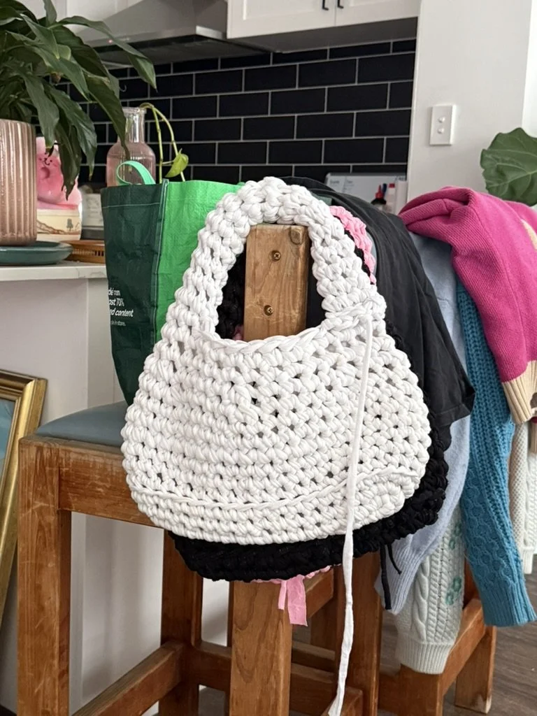 A white and black chunky knit handbag hanging on the back of a wooden chair in a cluttered room, with various clothing items and plants in the background.