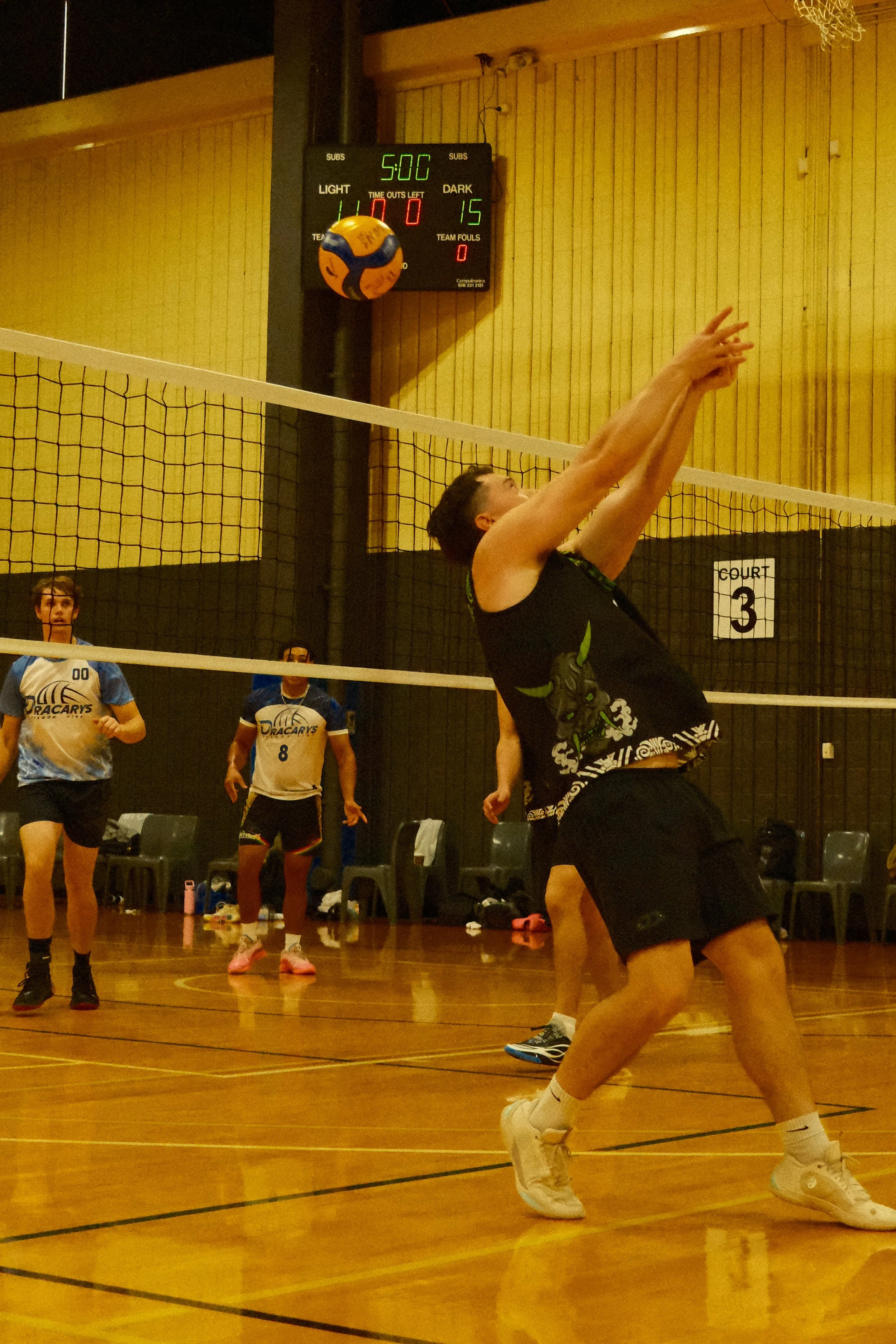 A volleyball game is in progress indoors. A player in black shorts and a black shirt is about to make a play at the net, with his arms extended upward. Other players are standing on the court, watching or preparing for the next move. The scoreboard shows 5:00 minutes remaining and 15 points for the dark team. The court has a wooden floor, and a volleyball net separates the teams. Some spectators are sitting on chairs along the court.