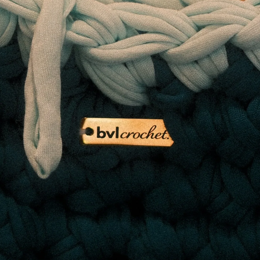 Close-up of a black quilted fabric with a beige fabric tag labeled 'bvlcrochet' attached, and grey woven laces tied atop the fabric.