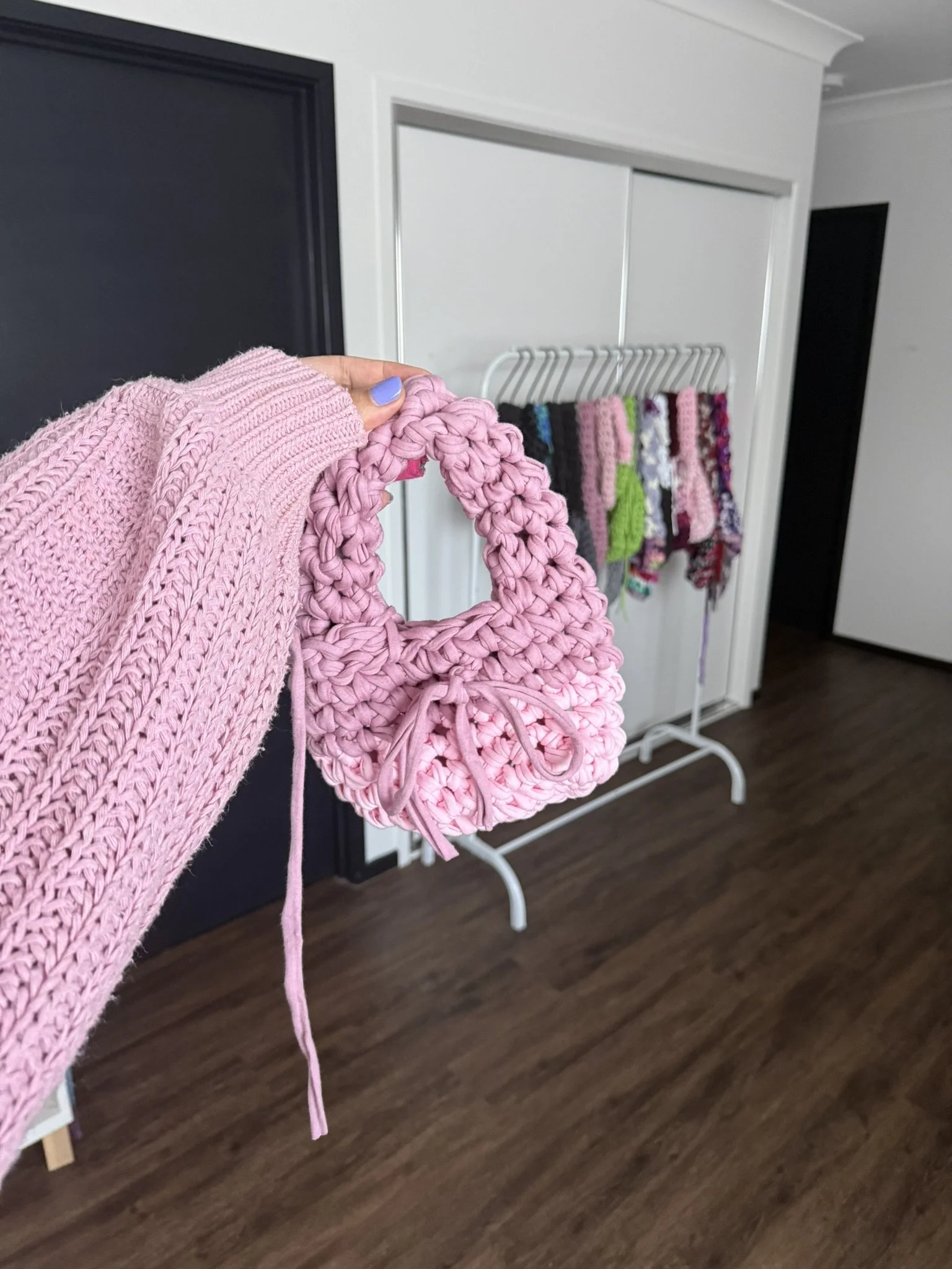 A person holding a pink woven handbag and a pink sweater in front of a white wall with a black door and a white clothing rack with colorful knit and fabric items.