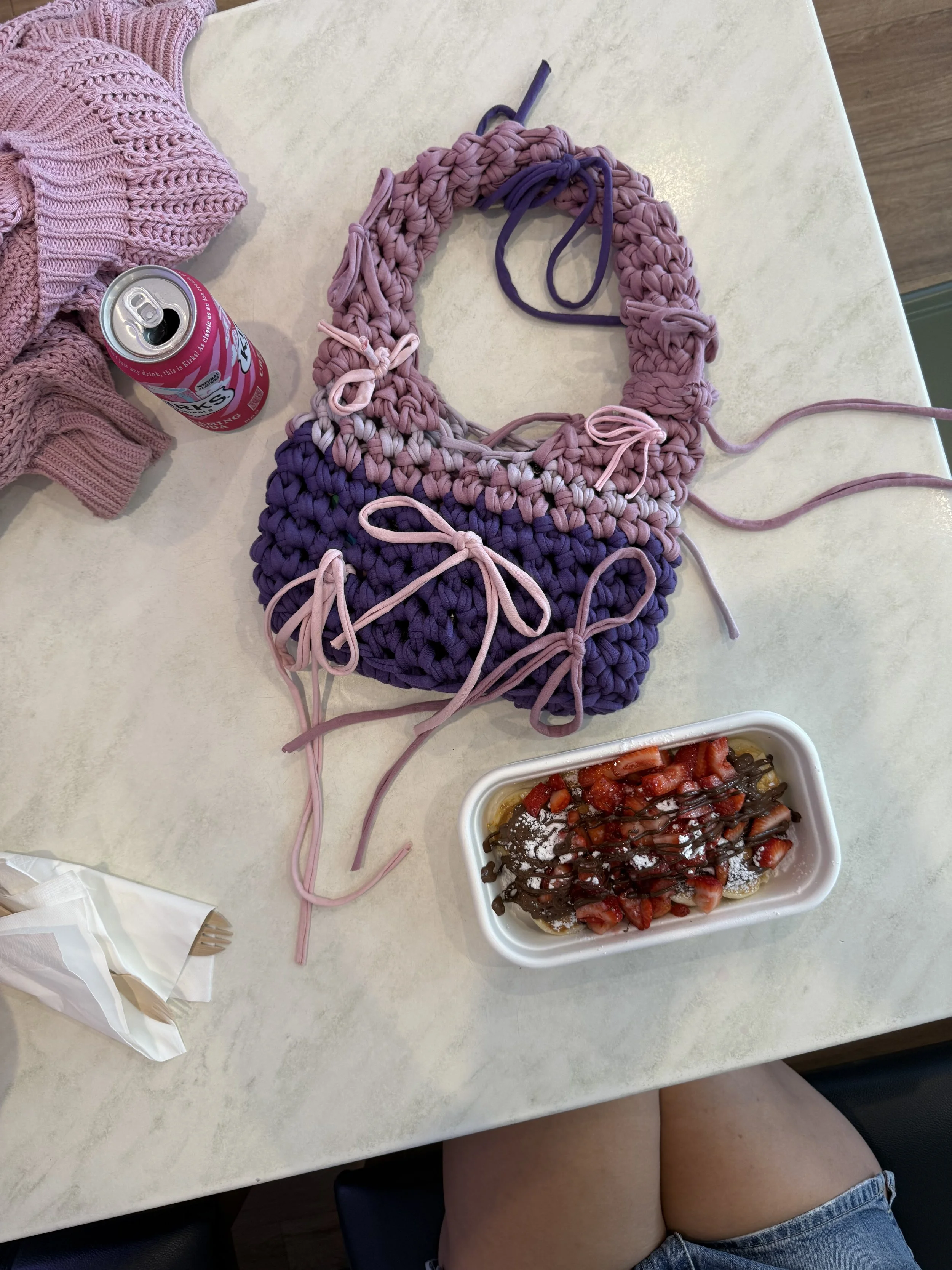 A table with a partially completed pink and purple chunky knit project, a pink knit garment, a can of soda, a paper napkin, and a container of strawberries and bananas with chocolate drizzle.