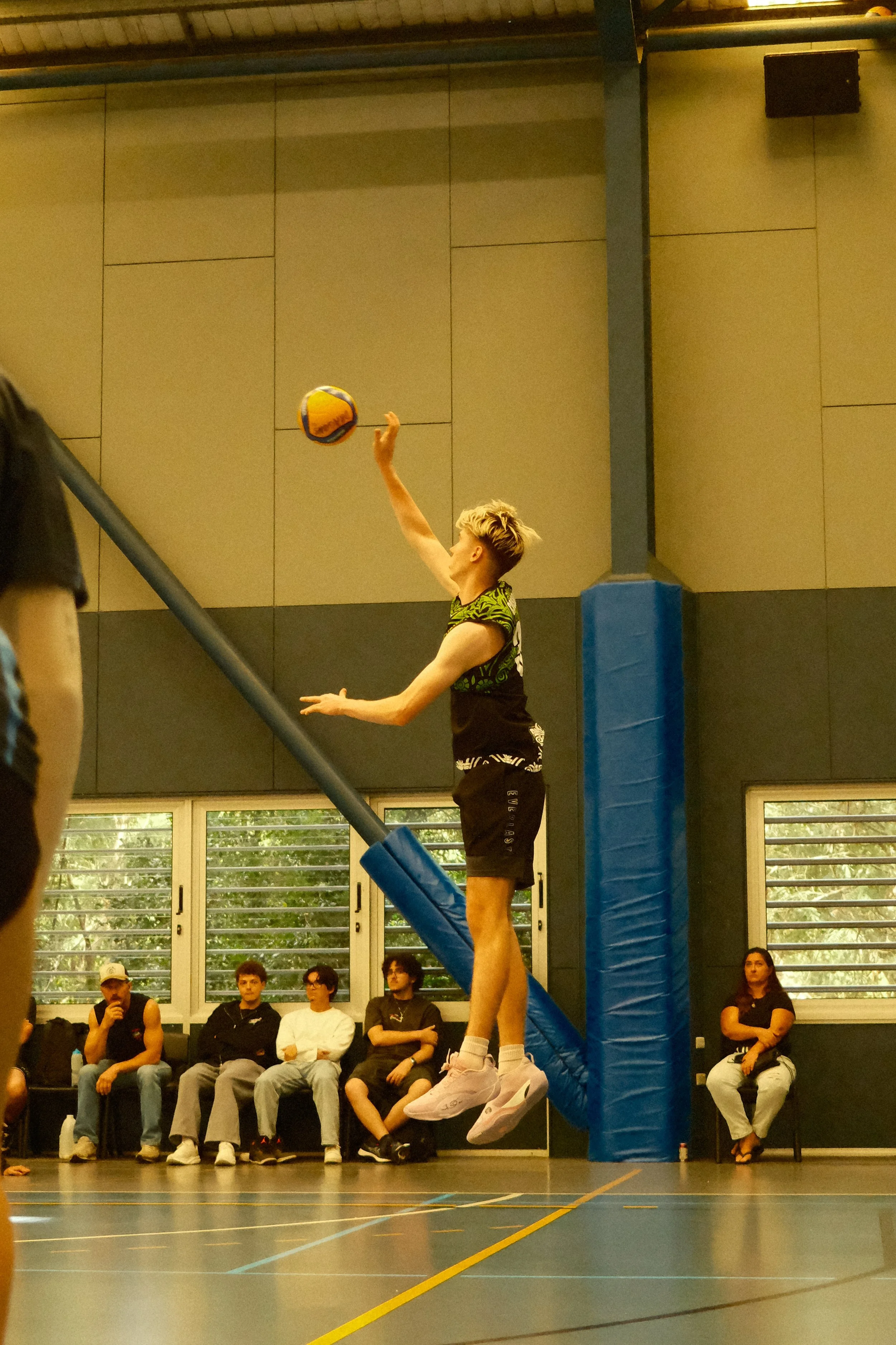 A volleyball player with blonde hair and black sportswear jumps to hit a volleyball during an indoor game, with spectators sitting and watching in the background.