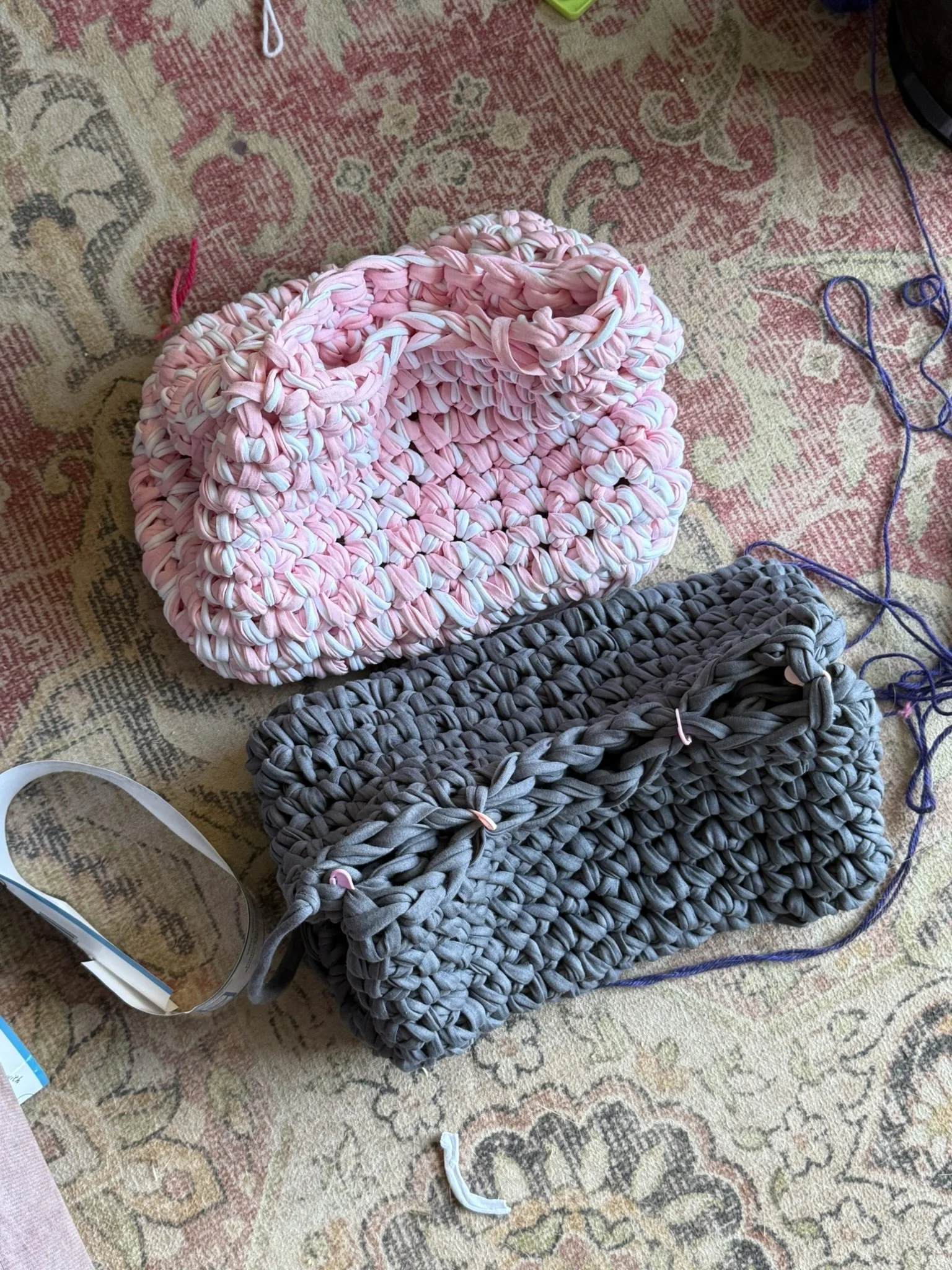 Two handmade crochet purses, one pink and white with a braided handle, and one dark gray with a similar braided handle, resting on a patterned area rug. There are strands of yarn and a clear plastic measuring tape nearby.