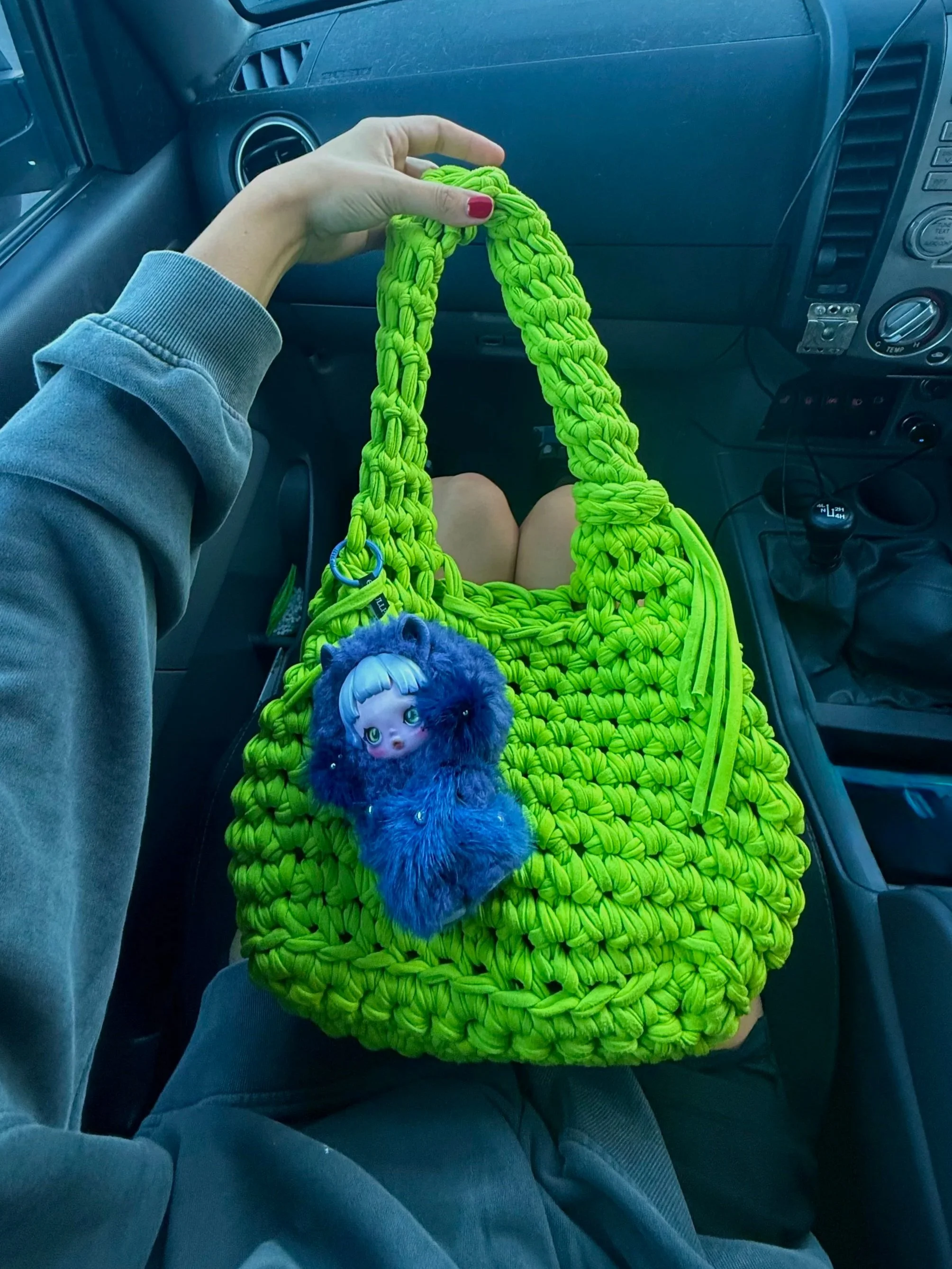 Person holding a bright yellow-green woven bag with a plush toy attached, sitting inside a vehicle with dashboard and gear shift visible.