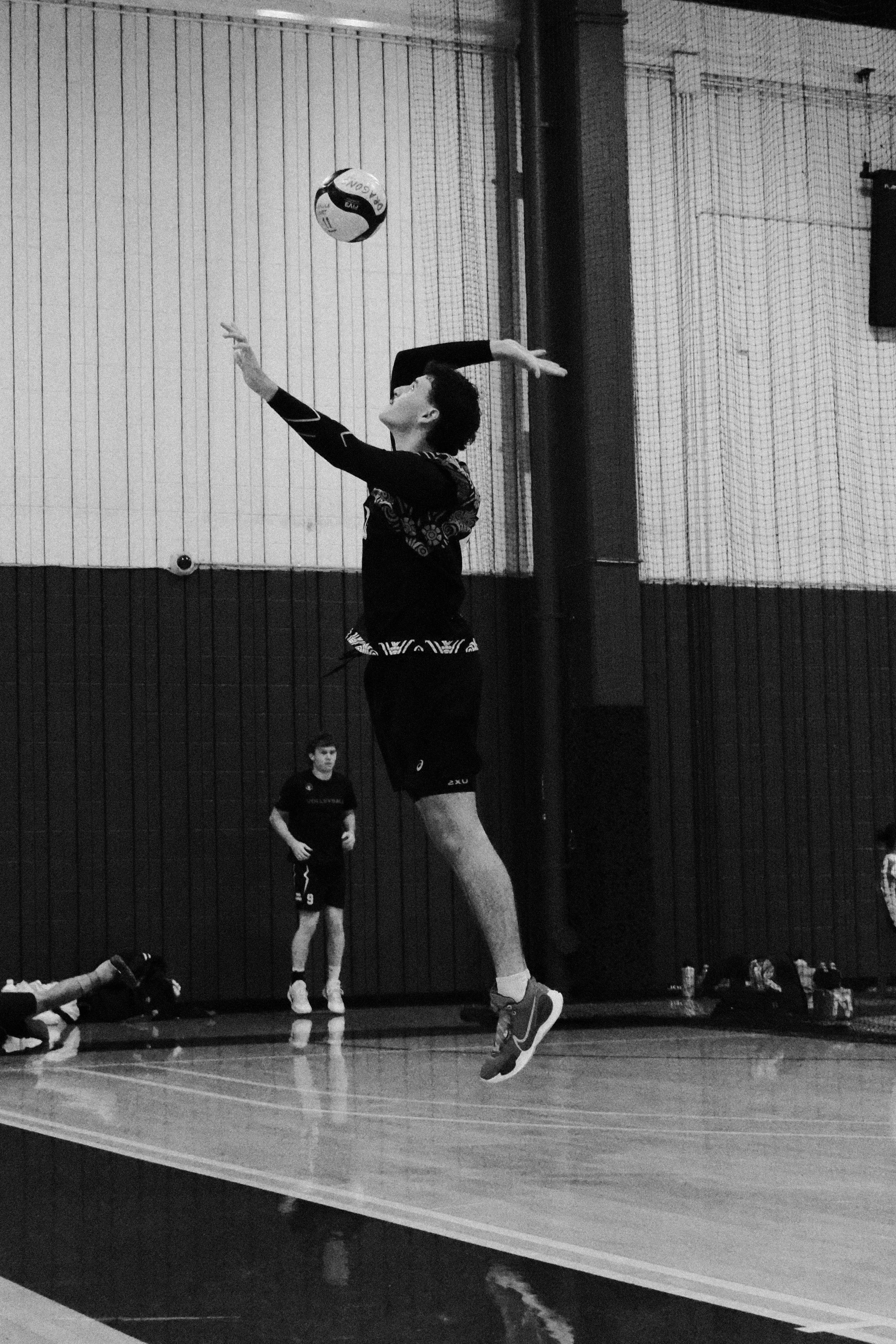 A person jumping to hit a volleyball during indoor volleyball practice, with others watching in the background and sports water bottles on the side.