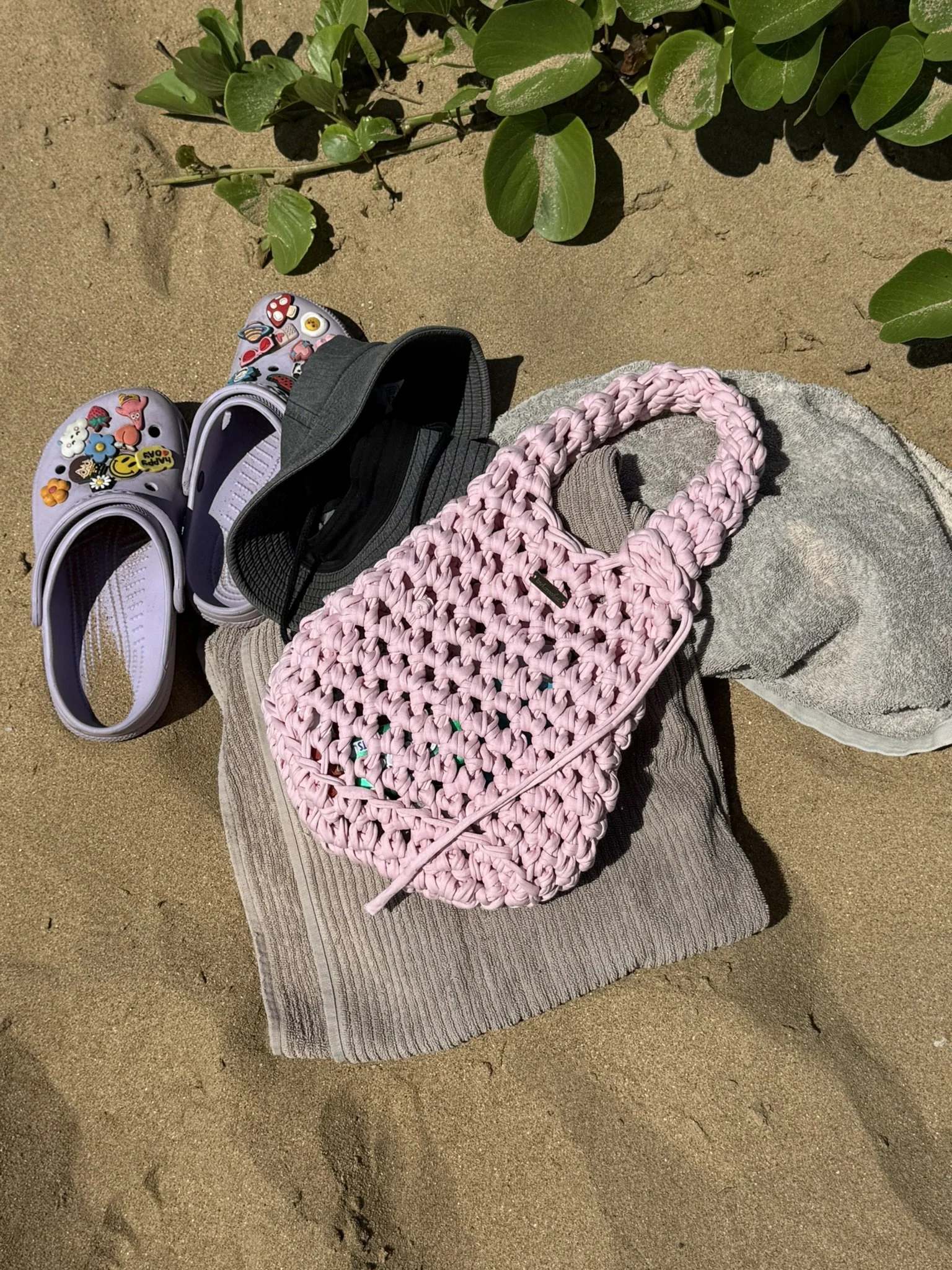A pair of purple Crocs decorated with colorful stickers, a black cap, a pink woven bag, a gray blanket, and a gray towel set on a brown striped mat on sandy ground, next to green leafy plants.
