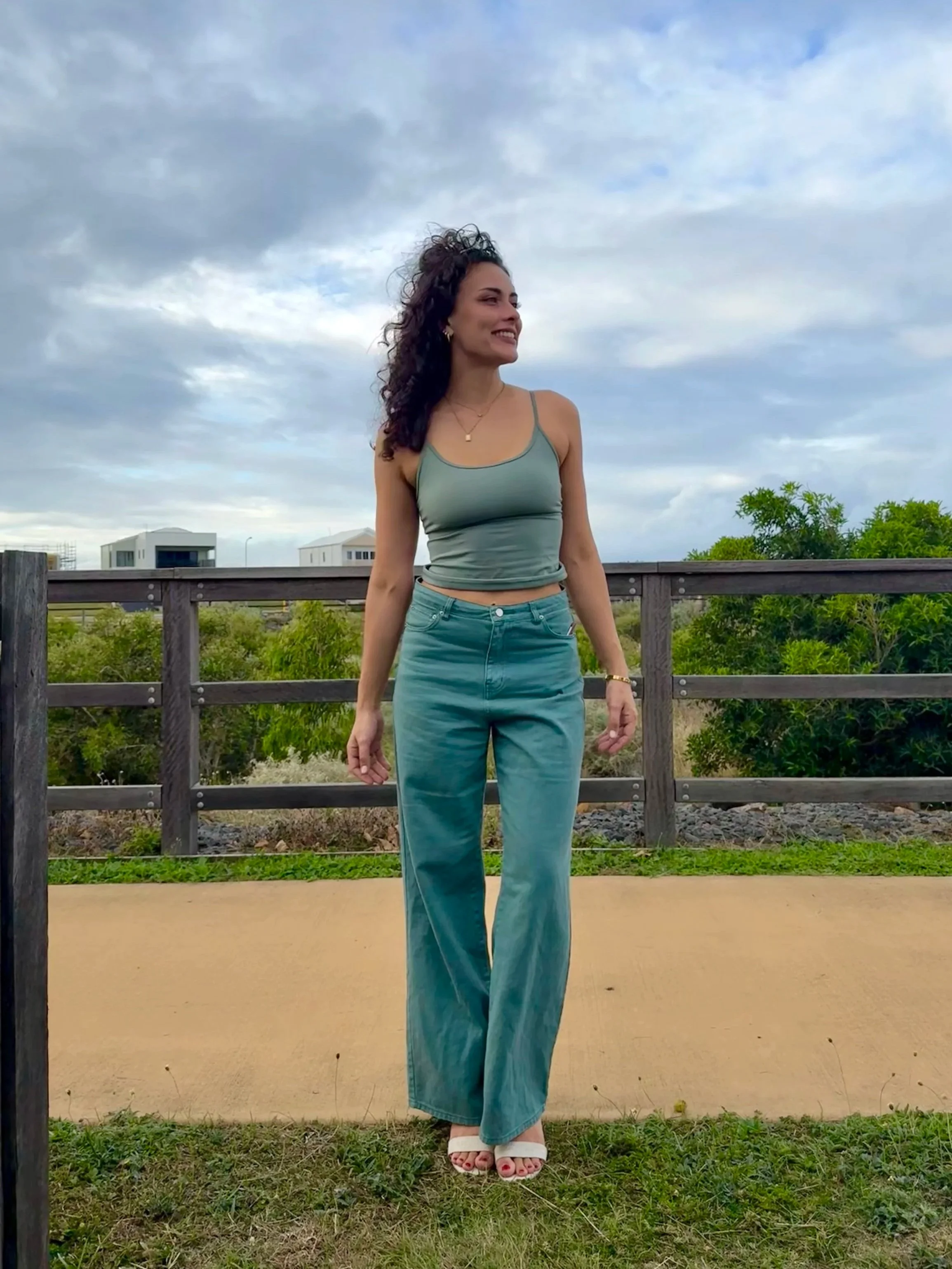 A woman with curly dark hair, wearing a gray tank top and high-waisted jeans, standing outdoors on a balcony or terrace with trees and modern houses in the background. She is smiling and looking to her left.