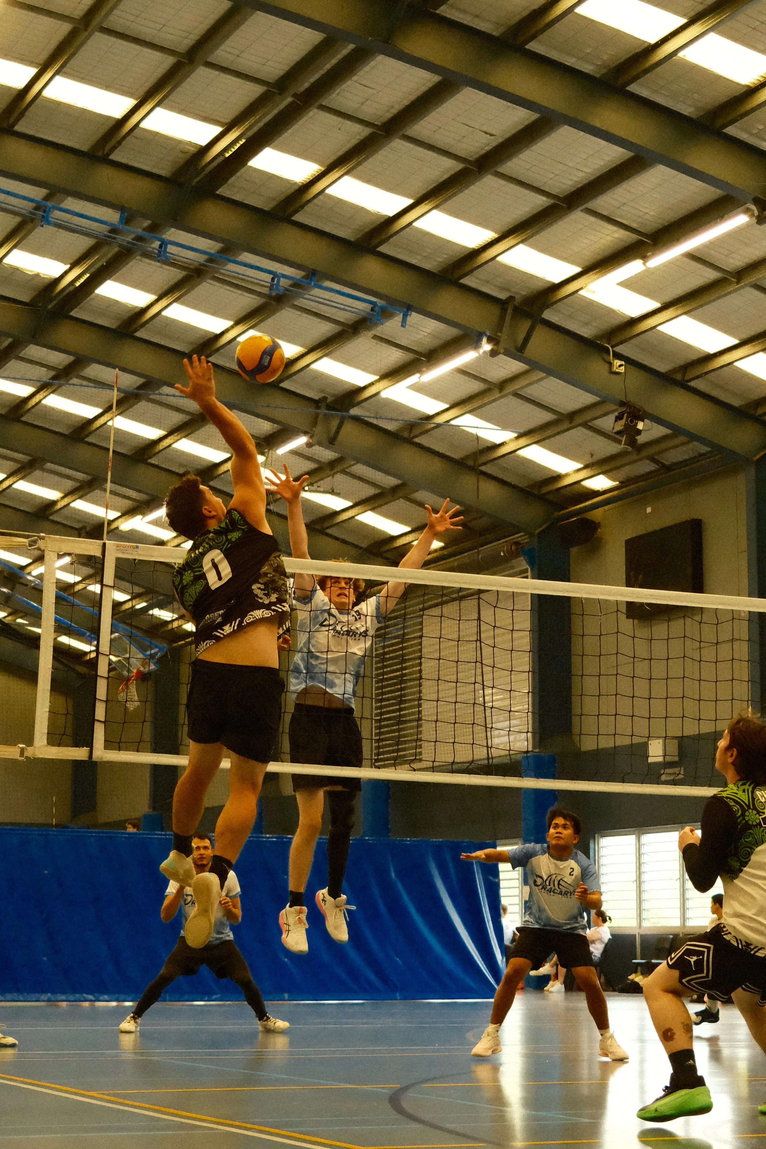 Indoor volleyball game with players jumping to block and hit the ball at the net, with spectators and other players in the background.